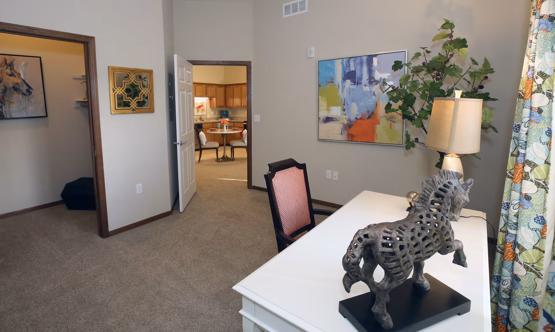 Interior view of a room with a white desk featuring a horse sculpture and a lamp. A chair with a red patterned backrest is positioned at the desk. The room has beige walls with colorful abstract artwork and a leafy plant near the window with floral curtains. An open door reveals a kitchen area with wooden cabinets and a small dining table with chairs.
