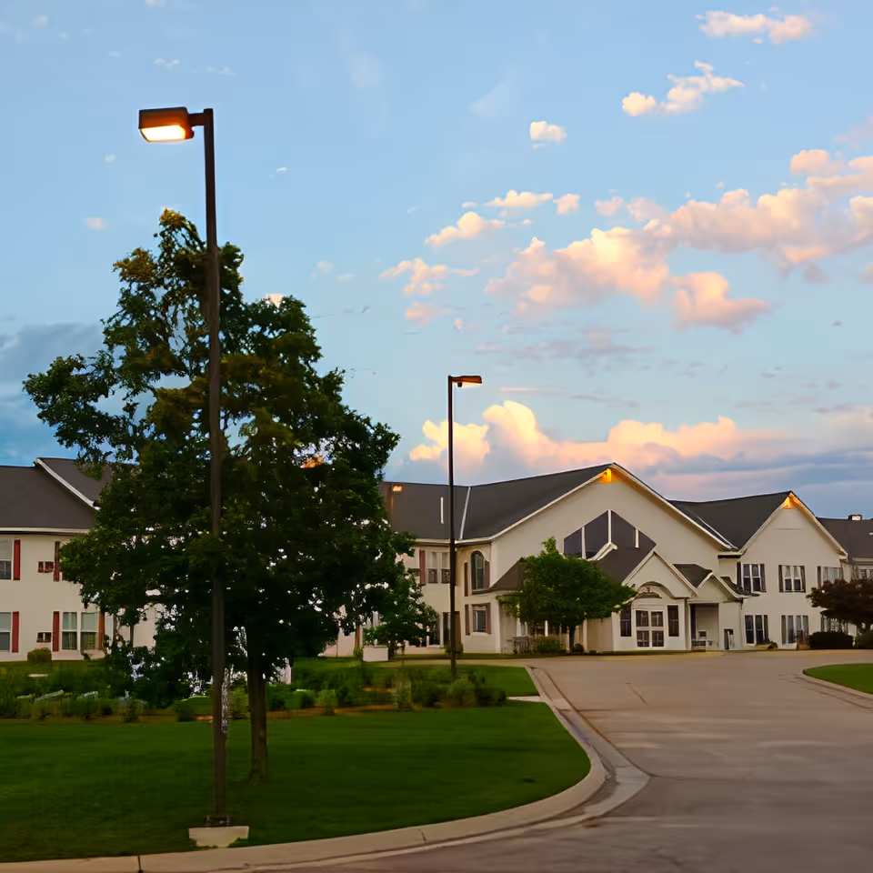 Exterior view of a senior living facility building with a driveway, green lawn, trees, and street lamps under a partly cloudy sky during sunset.