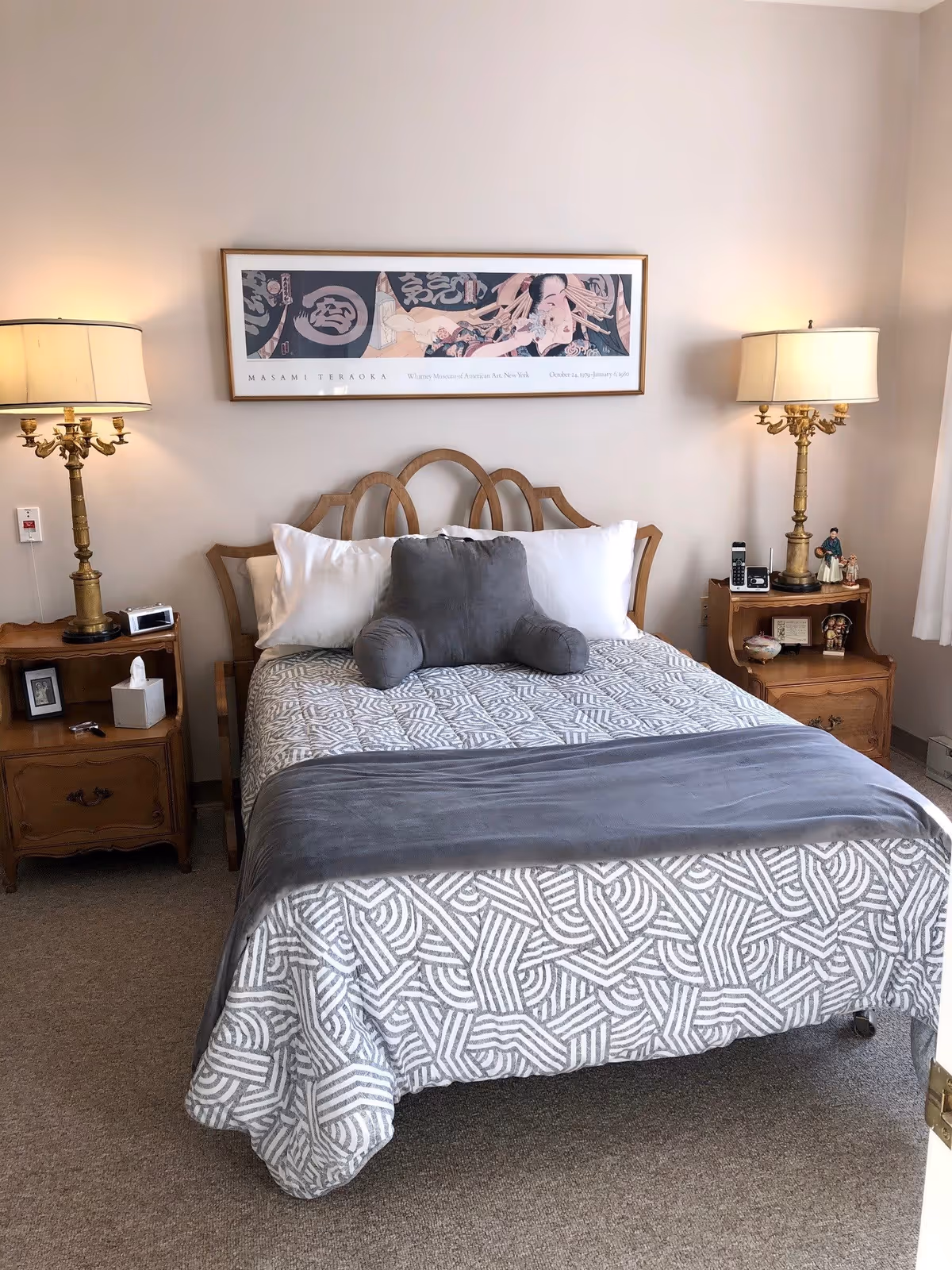 A neatly made bed with a patterned gray and white bedspread and a gray armrest pillow in the center. The bed has a wooden headboard and is flanked by two wooden nightstands, each with a brass lamp with a cream-colored shade. On the left nightstand, there is a tissue box, a small framed picture, and a clock. On the right nightstand, there are a few decorative items and a cordless phone. Above the bed hangs a framed artwork with Japanese characters and a reclining figure. The room has beige walls and carpeted flooring.
