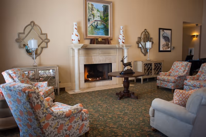 A cozy living room with a lit fireplace, floral patterned armchairs, a round wooden table with a deer sculpture, decorative mirrors, vases, and framed artwork on beige walls.