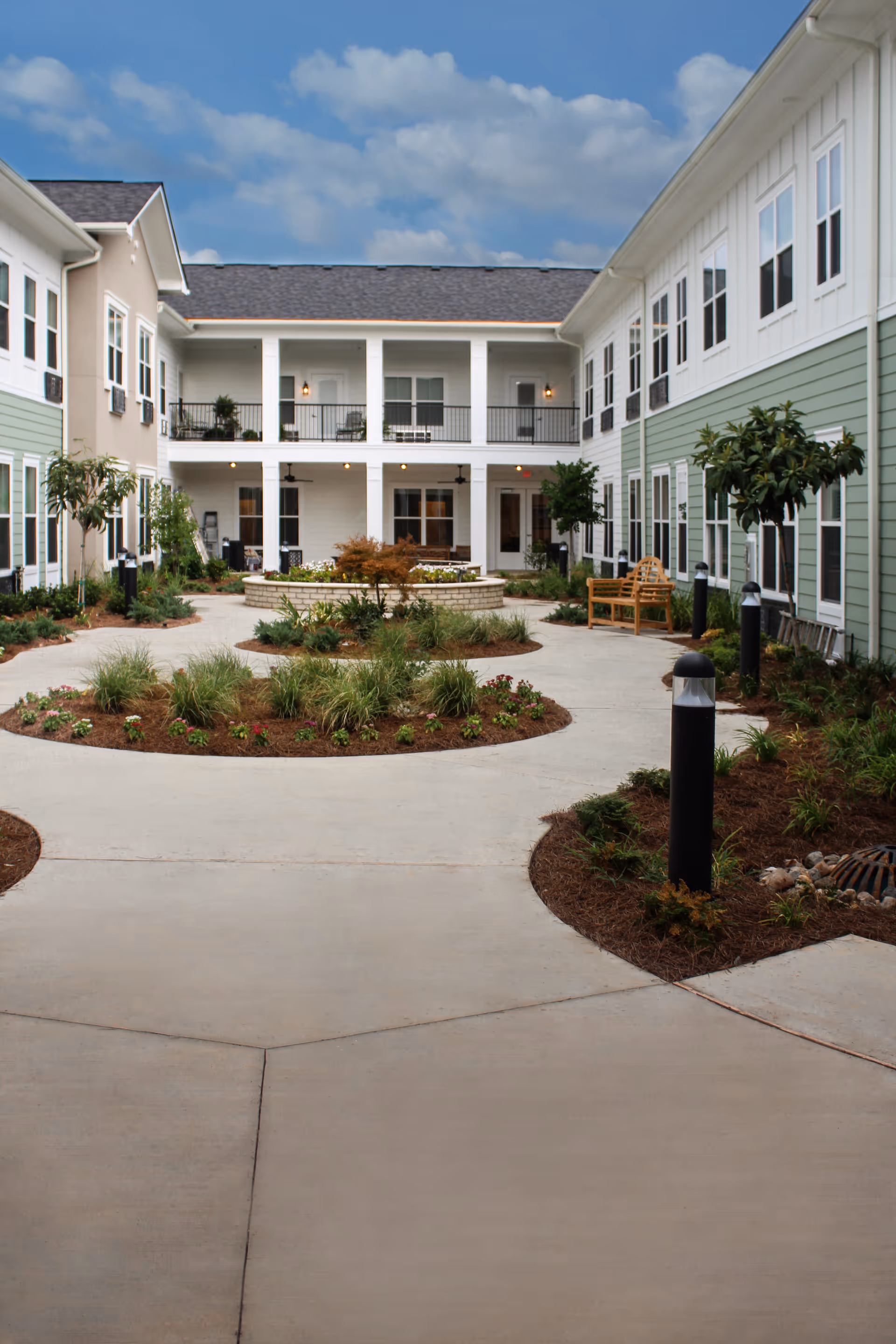 Outdoor courtyard area of The Blake at Bossier City senior living facility with a circular garden bed in the center, surrounded by a concrete walkway, benches, small trees, and two-story building with balconies and multiple windows under a partly cloudy sky.