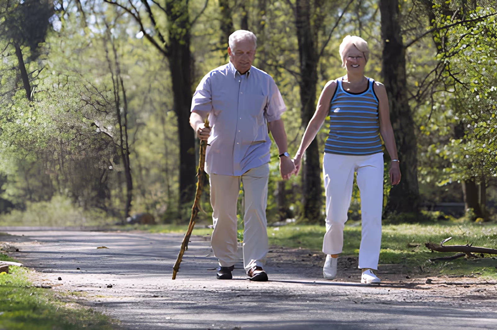 An elderly couple walking hand in hand on a paved path in a wooded park area. The man is using a walking stick and wearing a light-colored shirt and pants, while the woman is dressed in a striped sleeveless top and white pants. Trees and greenery surround the path on both sides.