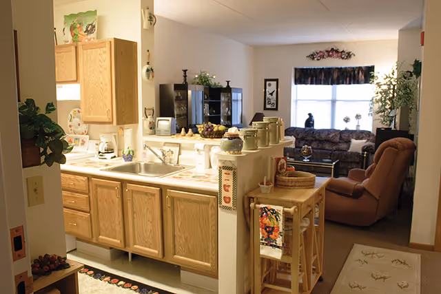 View of a cozy kitchen area with wooden cabinets and a sink, opening into a living room with a brown recliner chair, a patterned sofa, a coffee table, and a large window with floral curtains. The kitchen counter has various jars and decorative items, and there are plants and framed pictures on the walls.