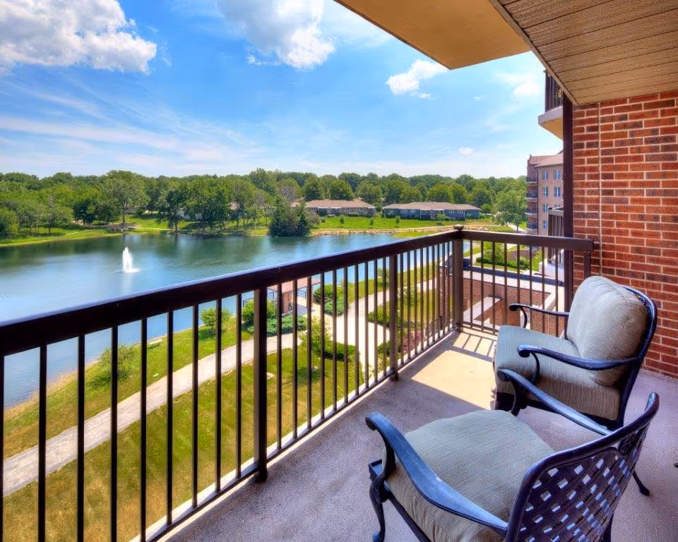 Balcony with two cushioned chairs overlooking a lake with a fountain and treed shoreline.