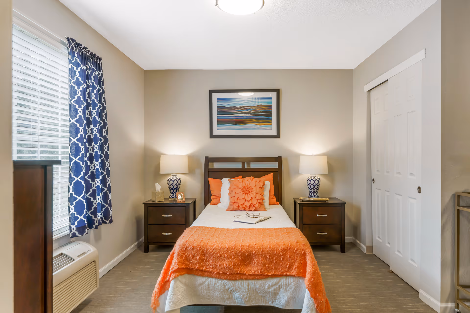 A neatly arranged bedroom with a single bed centered between two dark wooden nightstands, each with a blue and white patterned lamp. The bed has white bedding with an orange throw blanket and decorative pillows. A framed abstract landscape painting hangs above the bed. To the left, there is a window with blue patterned curtains and a wall-mounted air conditioning unit below it. On the right side, there is a white sliding closet door.