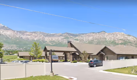 Exterior view of Quail Meadow Assisted Living facility with a parking lot in front, a beige fence, and mountains in the background under a clear blue sky.