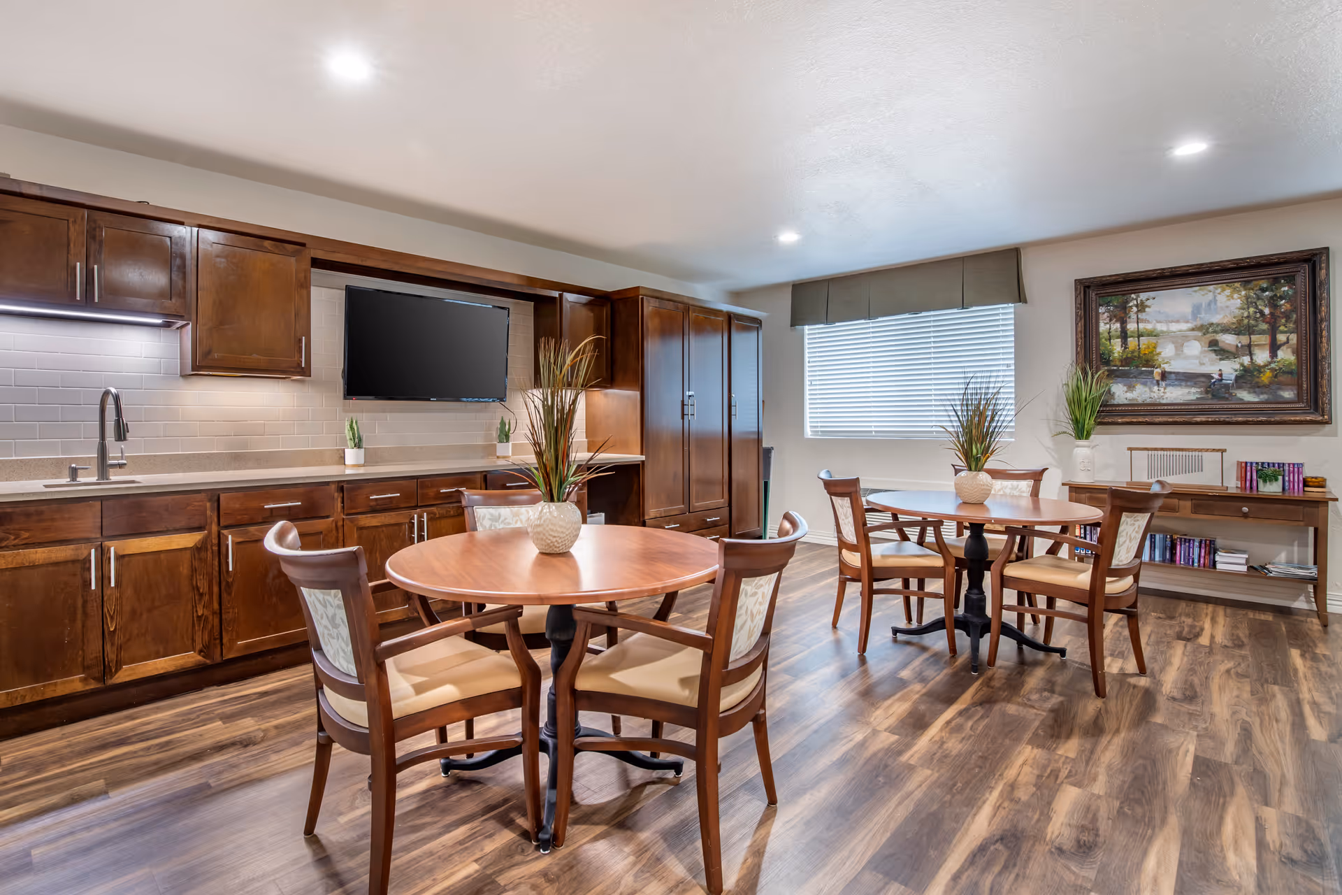 A well-lit room with two round wooden tables, each surrounded by four wooden chairs with cushioned seats. The room features wooden cabinetry along one wall with a mounted flat-screen TV, a countertop with a sink, and decorative plants. A large window with blinds and a valance lets in natural light. On the opposite wall, there is a framed painting above a console table with books and decorative items. The floor is covered with wood-patterned laminate.