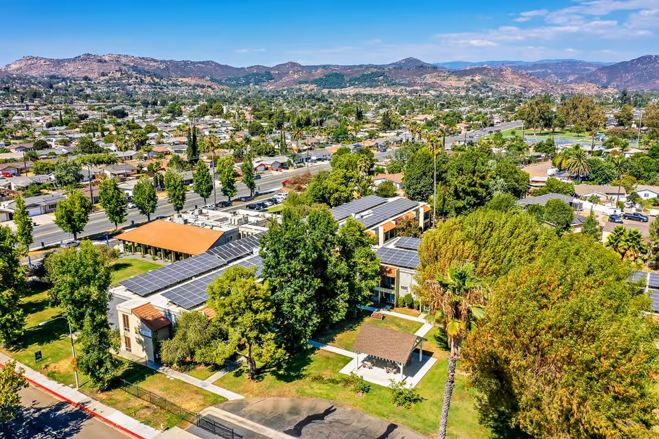 Aerial view of Escondido Senior Living facility showing multiple buildings with solar panels on the roofs, surrounded by green trees and lawns. The facility is located in a suburban area with many houses and roads visible, and mountains in the background under a partly cloudy sky.