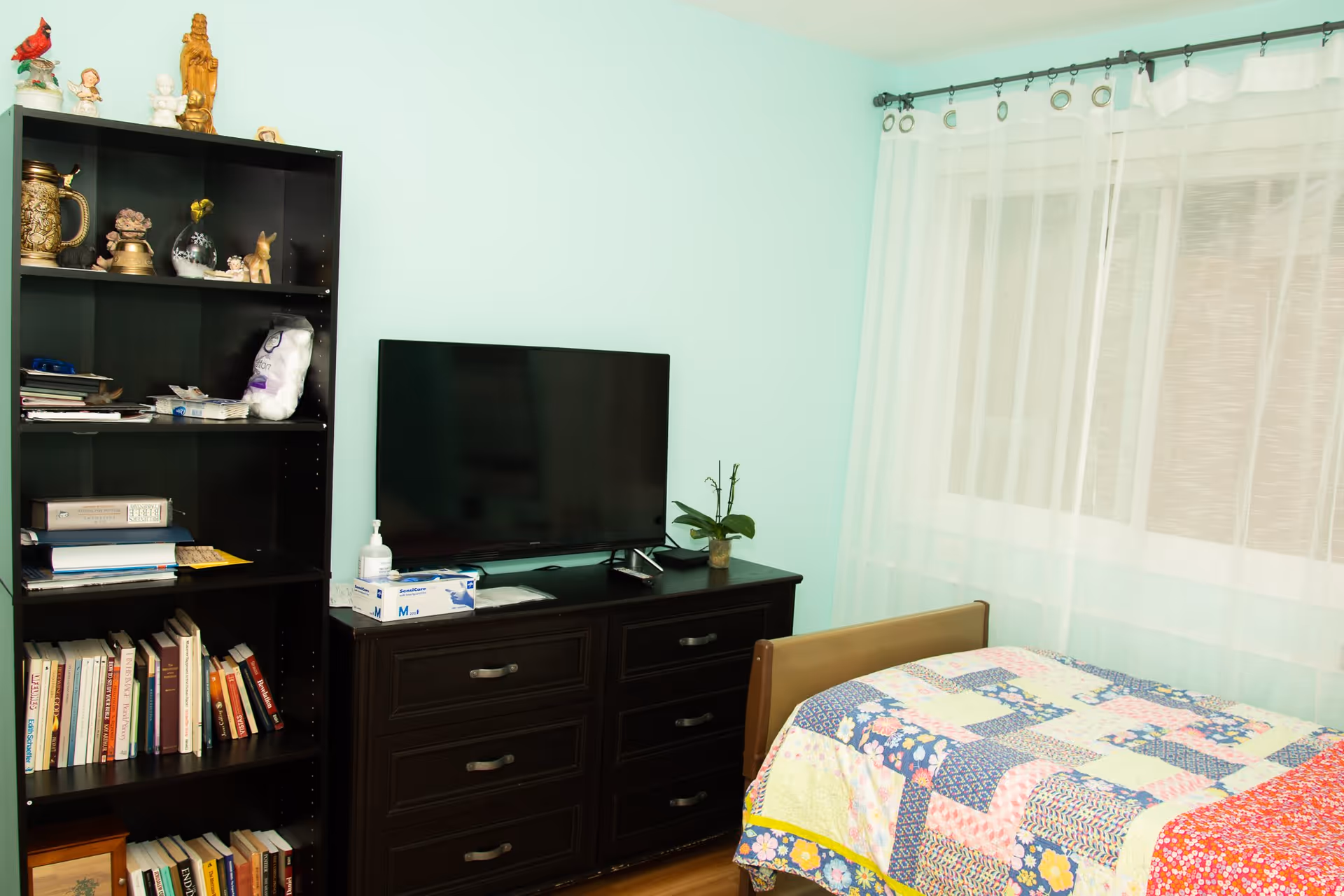 Small bedroom with a colorful patchwork quilted bed, a dresser topped with a TV and plant, and a bookshelf against a mint-green wall.