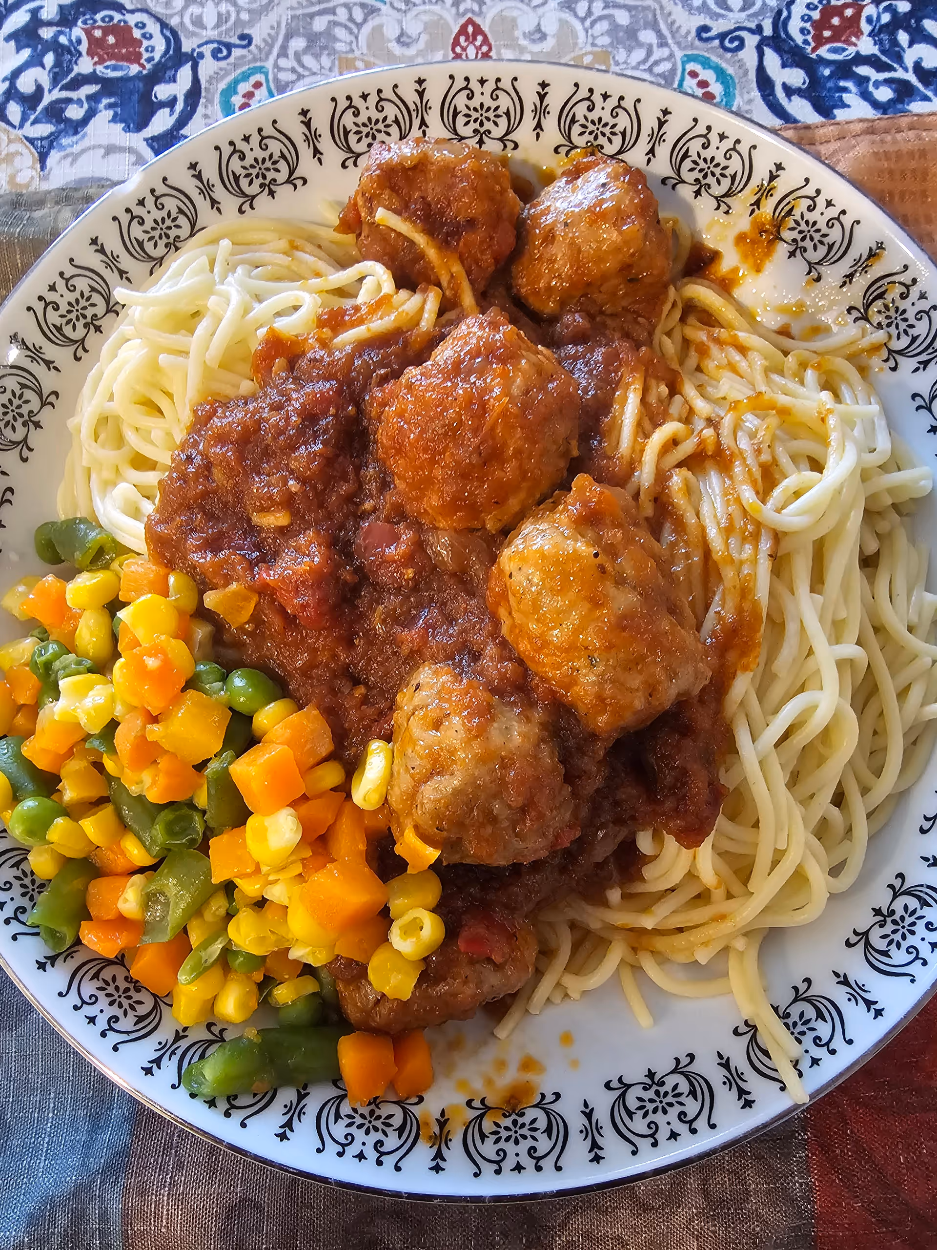 A plate of spaghetti topped with meatballs and tomato sauce, served with a side of mixed vegetables including corn, carrots, green beans, and peas. The plate has a decorative black and white pattern around the rim and is placed on a colorful tablecloth.