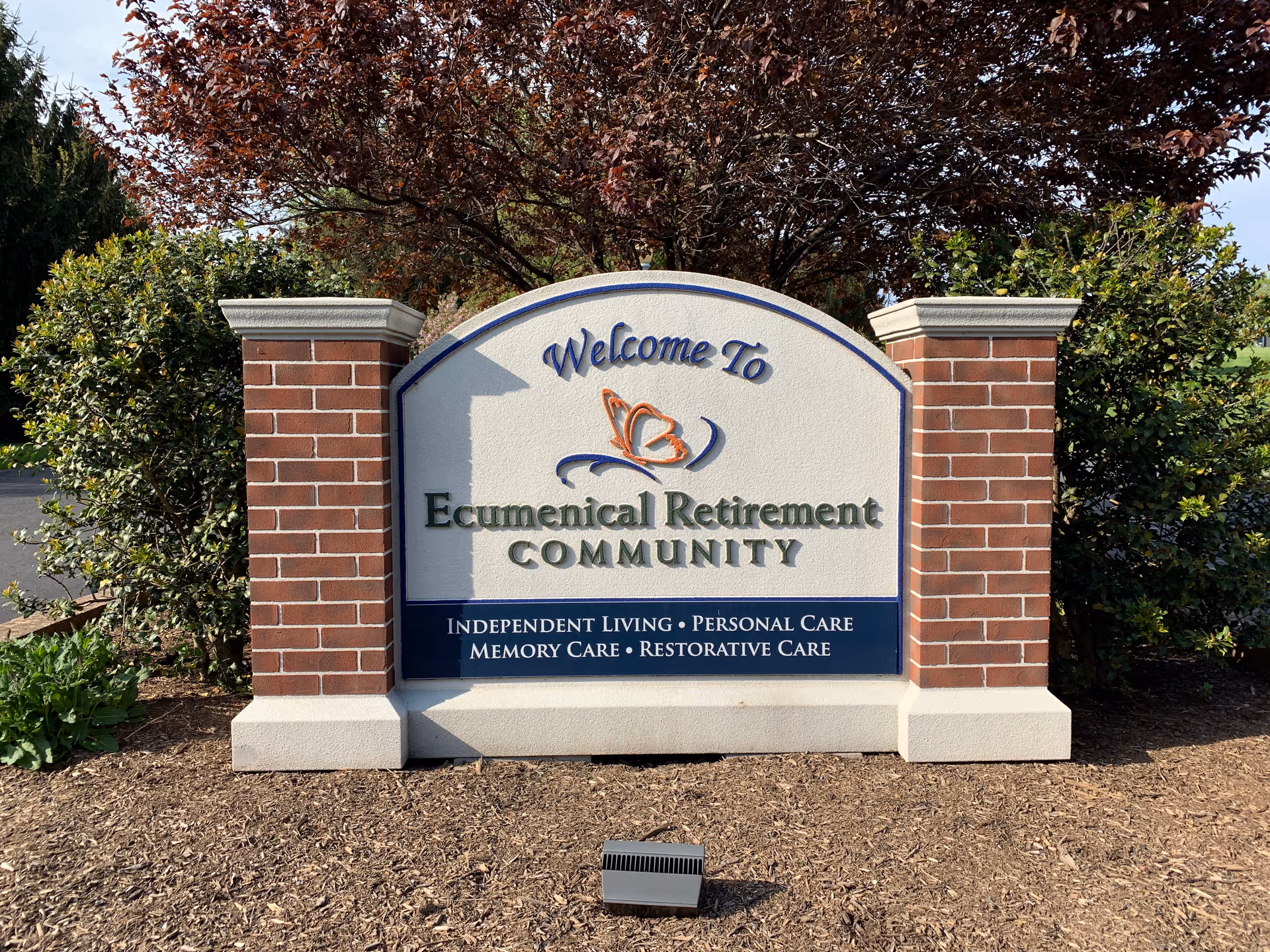 A welcoming sign for Ecumenical Retirement Community made of brick and stone, surrounded by bushes and trees with red and green leaves. The sign lists services including Independent Living, Personal Care, Memory Care, and Restorative Care.