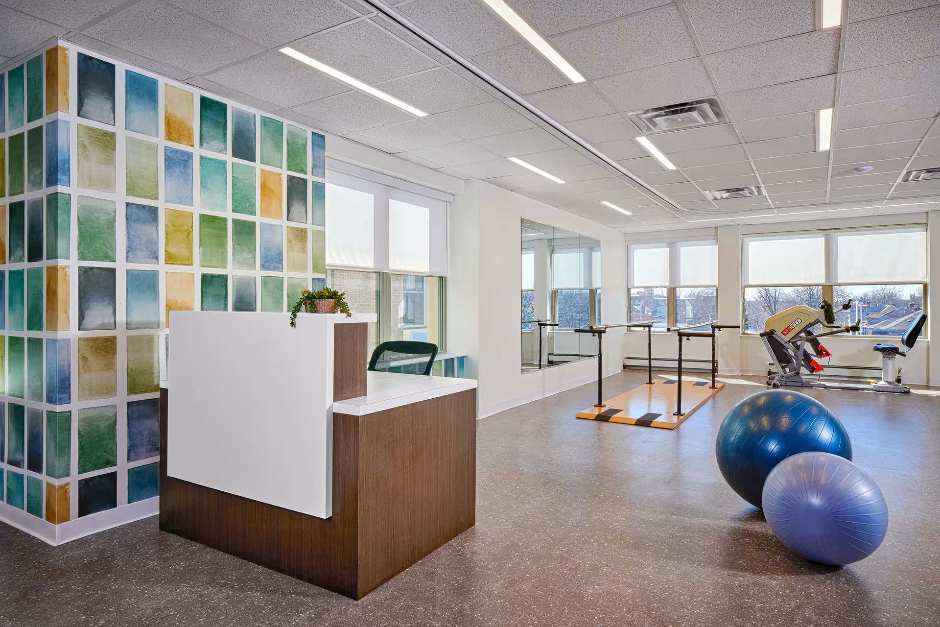 A bright and spacious exercise room with large windows letting in natural light. The room features a colorful tiled wall, a small reception desk with a plant on it, two exercise balls on the floor, ballet barres, a large mirror on the wall, and exercise equipment near the windows.
