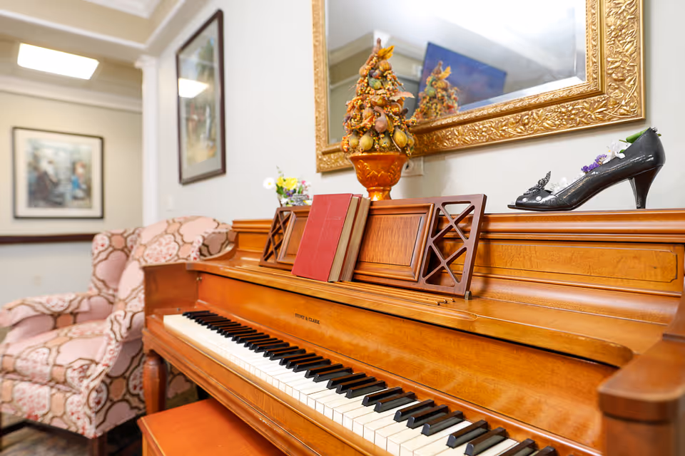 Upright wooden piano topped with books, decorative ornaments and a black high-heeled shoe, beside a patterned upholstered armchair in a sitting area.