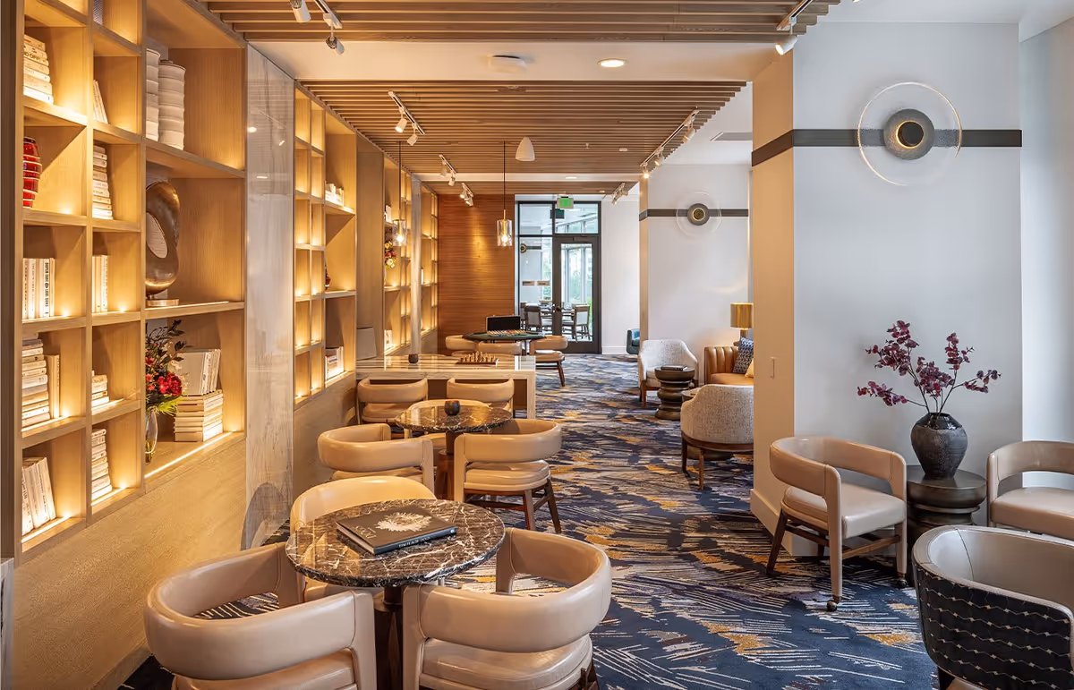 A well-lit, modern lounge area with multiple round tables and beige cushioned chairs arranged for small group seating. The left wall features built-in wooden shelves filled with books and decorative items, illuminated by warm lighting. The floor is covered with a patterned carpet in shades of blue and beige. The ceiling has wooden slats with track lighting. There are additional seating areas with armchairs and side tables along the right side, and a vase with purple flowers on a small table near the wall.
