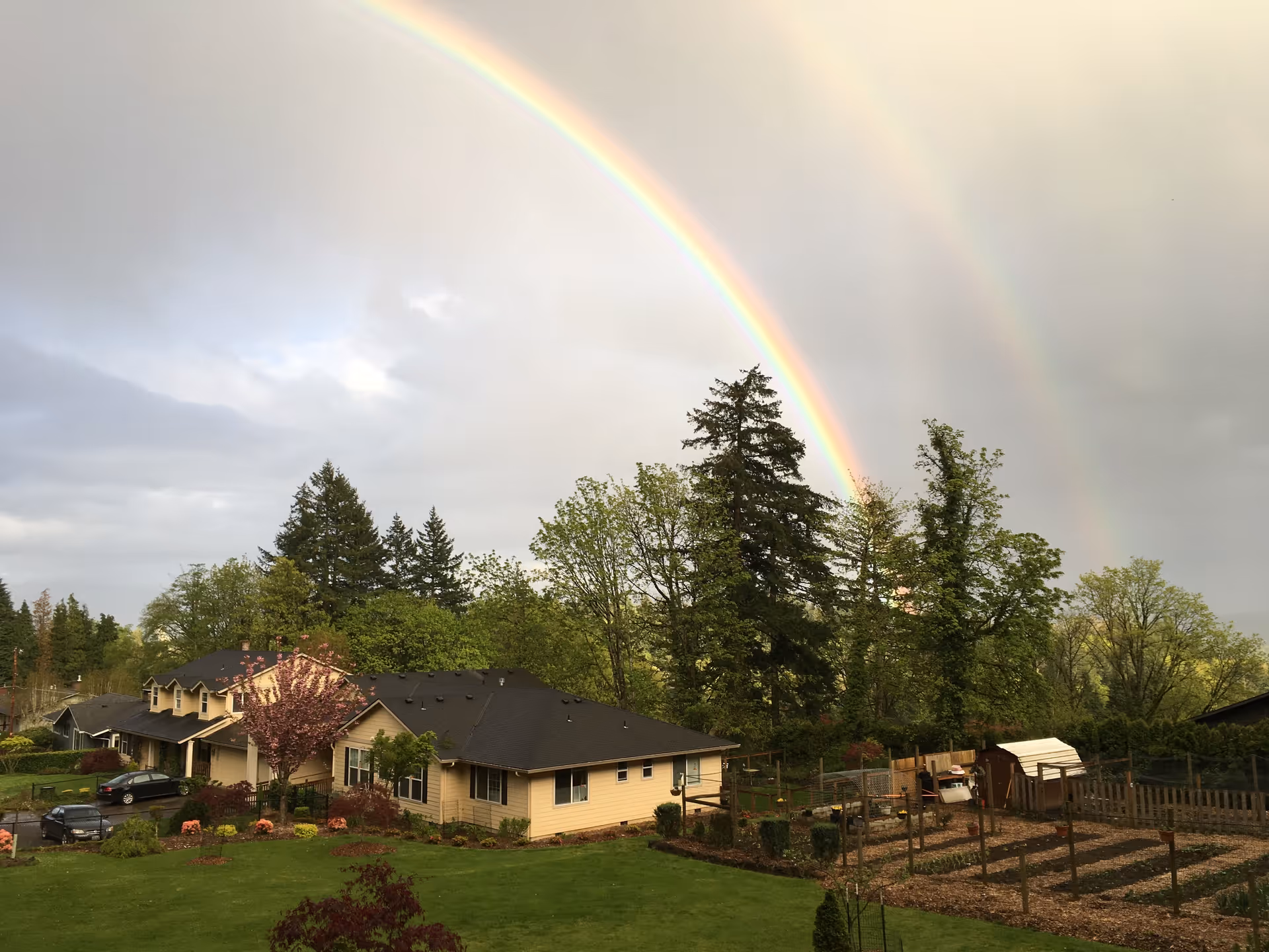 A scenic outdoor view of a residential facility with beige buildings, surrounded by green trees and a garden area. A vibrant double rainbow arcs across a cloudy sky above the property.
