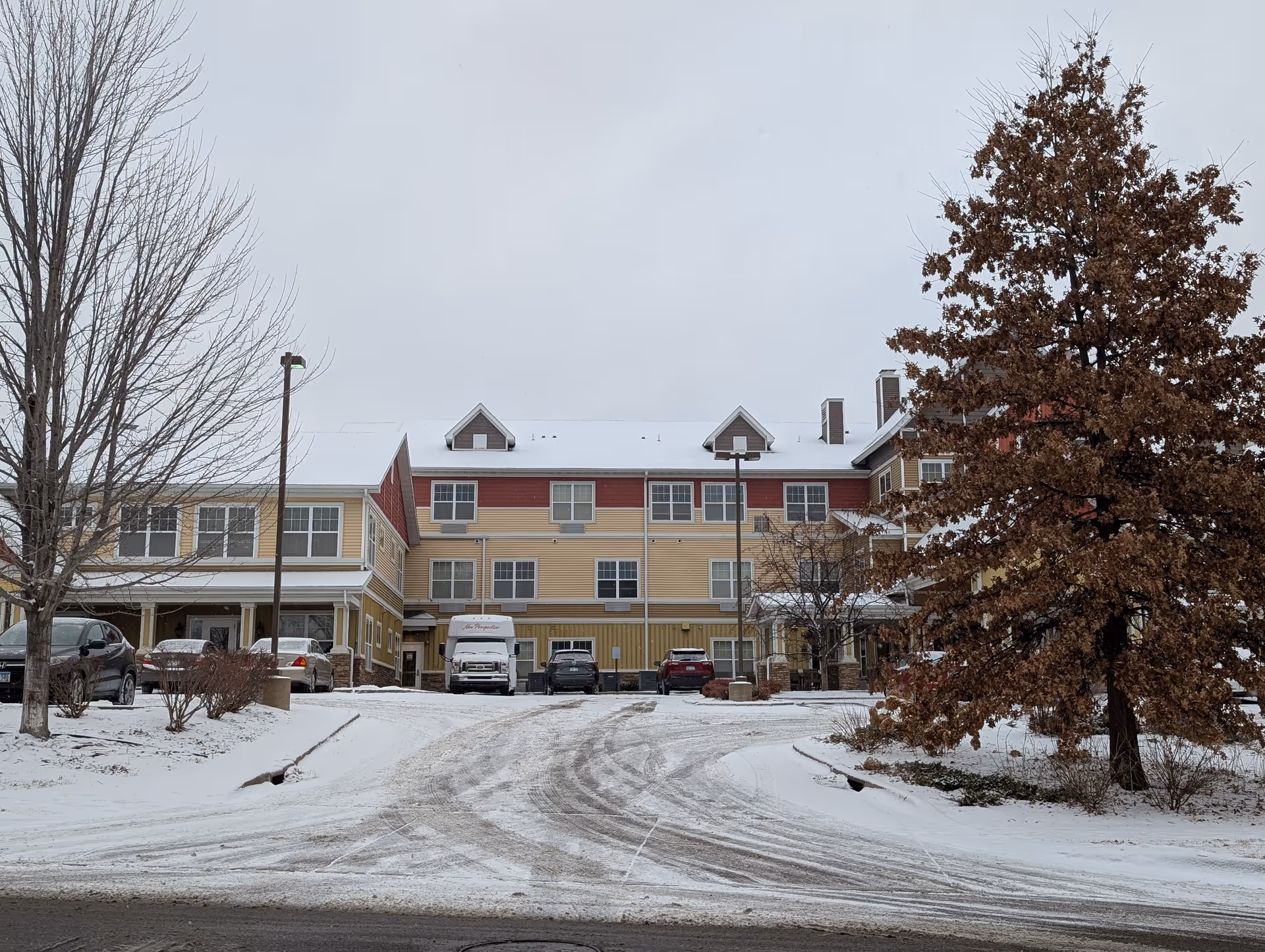 Front exterior of a three-story senior living building with a snow-covered driveway, parked cars, and bare trees.