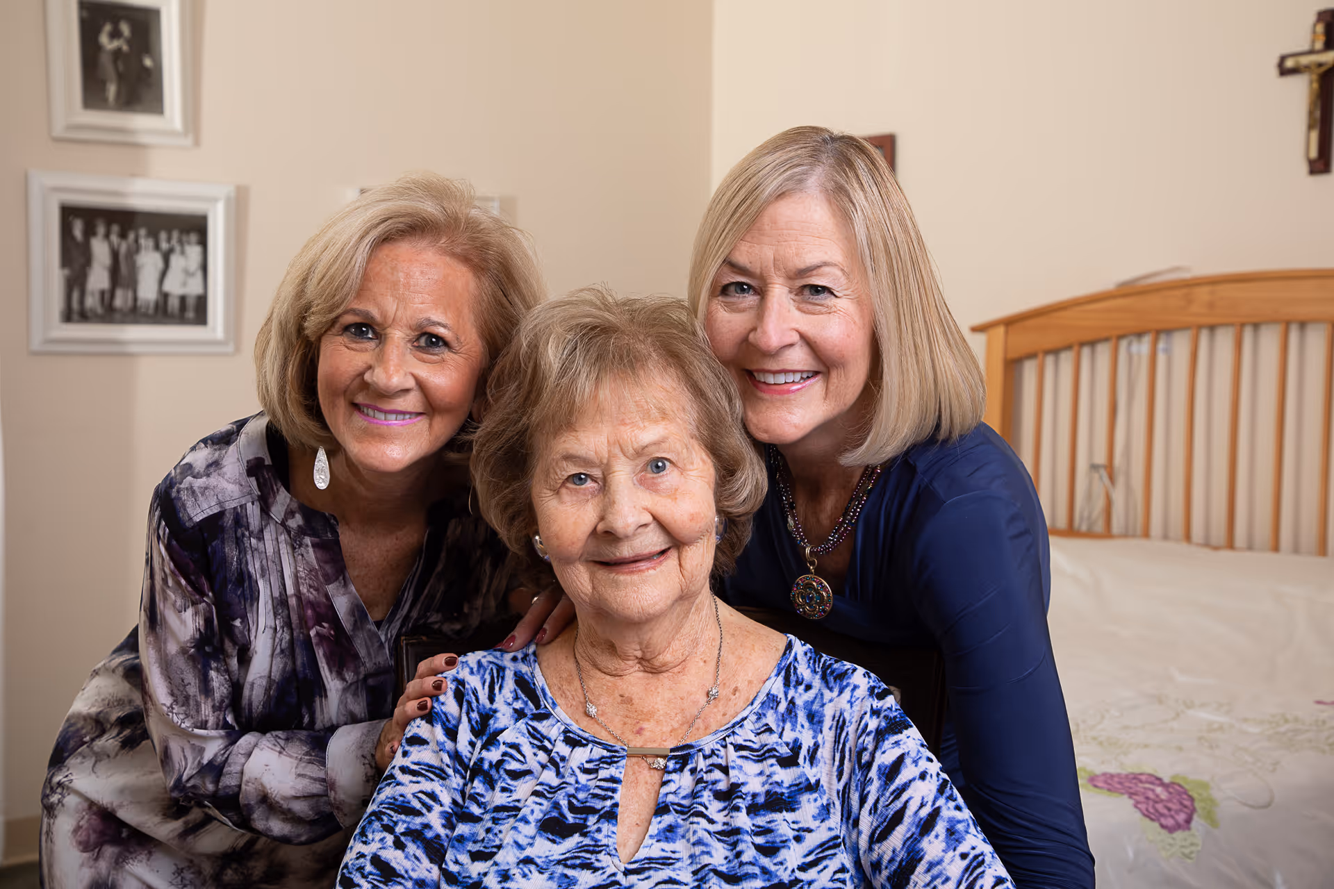 Three smiling women, two standing behind and one seated in front, in a bedroom setting with a wooden bed frame and framed photos on the wall.