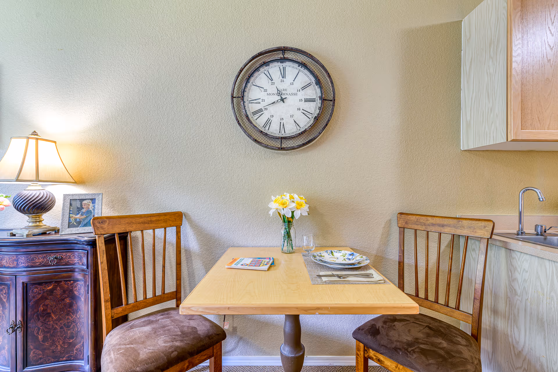 Small dining nook with a table set for two, wooden chairs, a wall clock, vase of flowers, and a lamp on a side cabinet next to a kitchenette sink.
