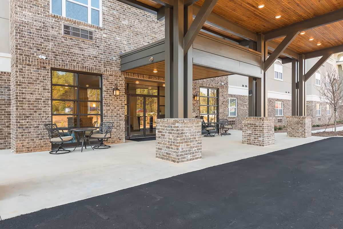 Covered entrance area of a senior living facility with brick pillars and walls, metal beams supporting a wooden ceiling, glass doors, and outdoor seating with metal chairs and tables.