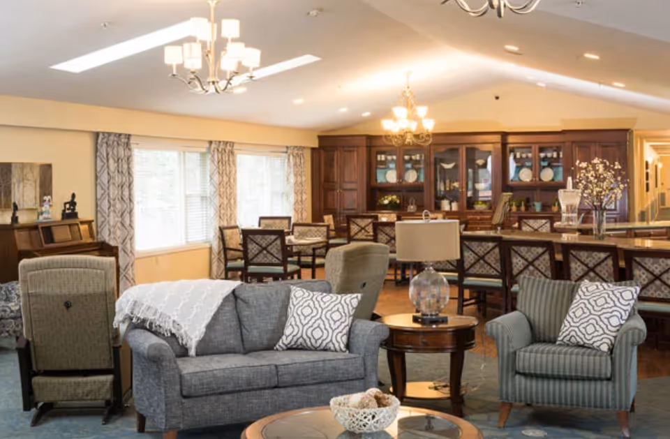 Bright communal living area with sofas, armchairs, a coffee table, and a dining area with wooden cabinets and chandeliers.