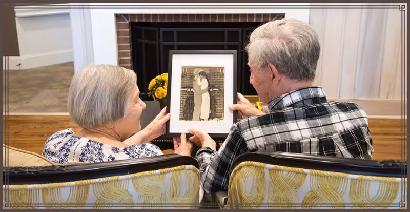 An elderly couple sitting on a couch in a living room, holding and looking at a framed black-and-white photograph of a couple embracing. The room has wooden flooring, a fireplace with yellow flowers on the mantel, and light-colored walls.