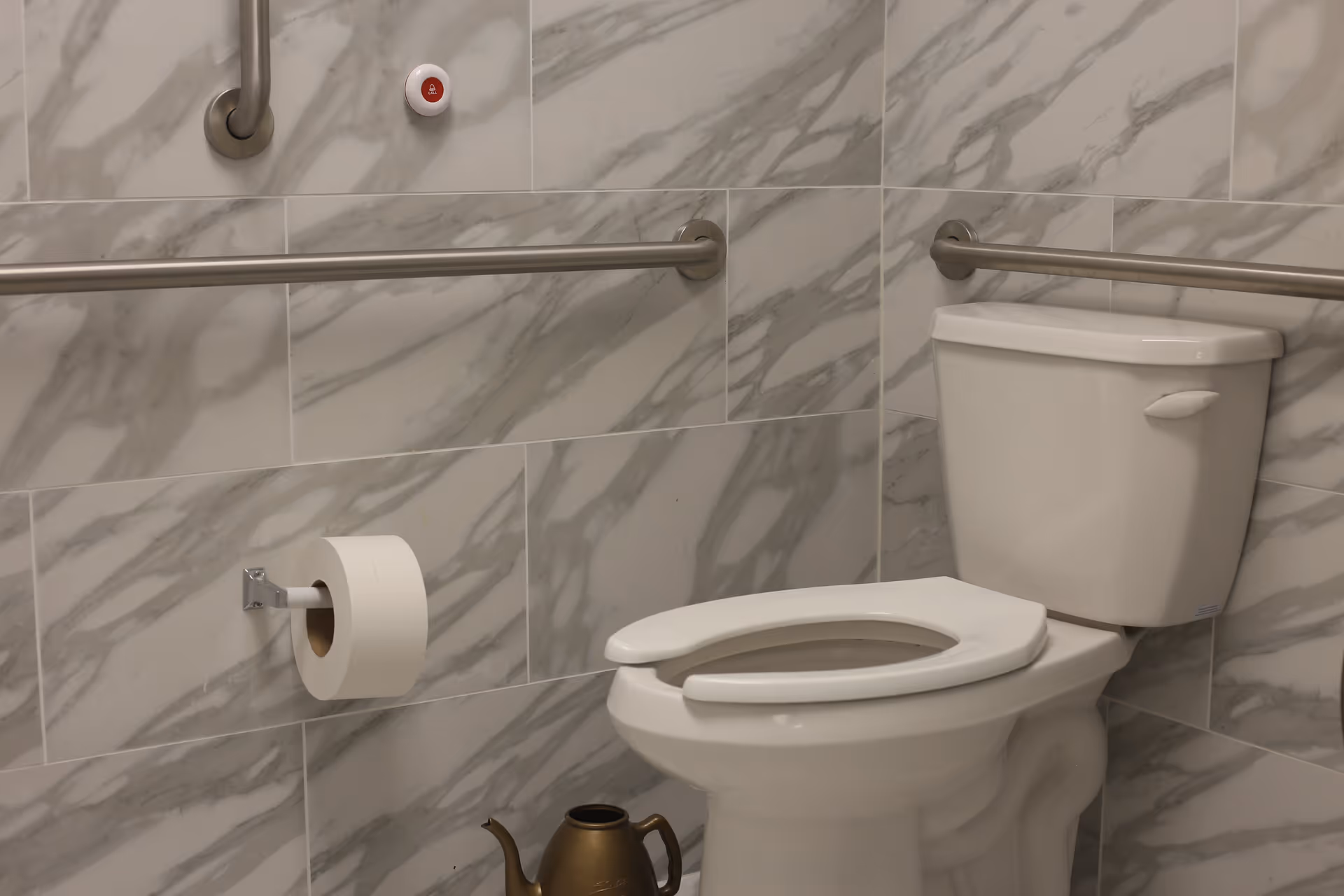 A clean, accessible bathroom with a white toilet featuring an open front seat, marble-patterned wall tiles, stainless steel grab bars on the walls, a toilet paper holder with a roll of toilet paper, and a bronze-colored watering can on the floor.