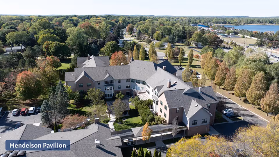 Aerial view of a large senior living campus with a multi-wing building labeled Mendelson Pavilion, surrounded by trees, parking lots, and a distant lake.