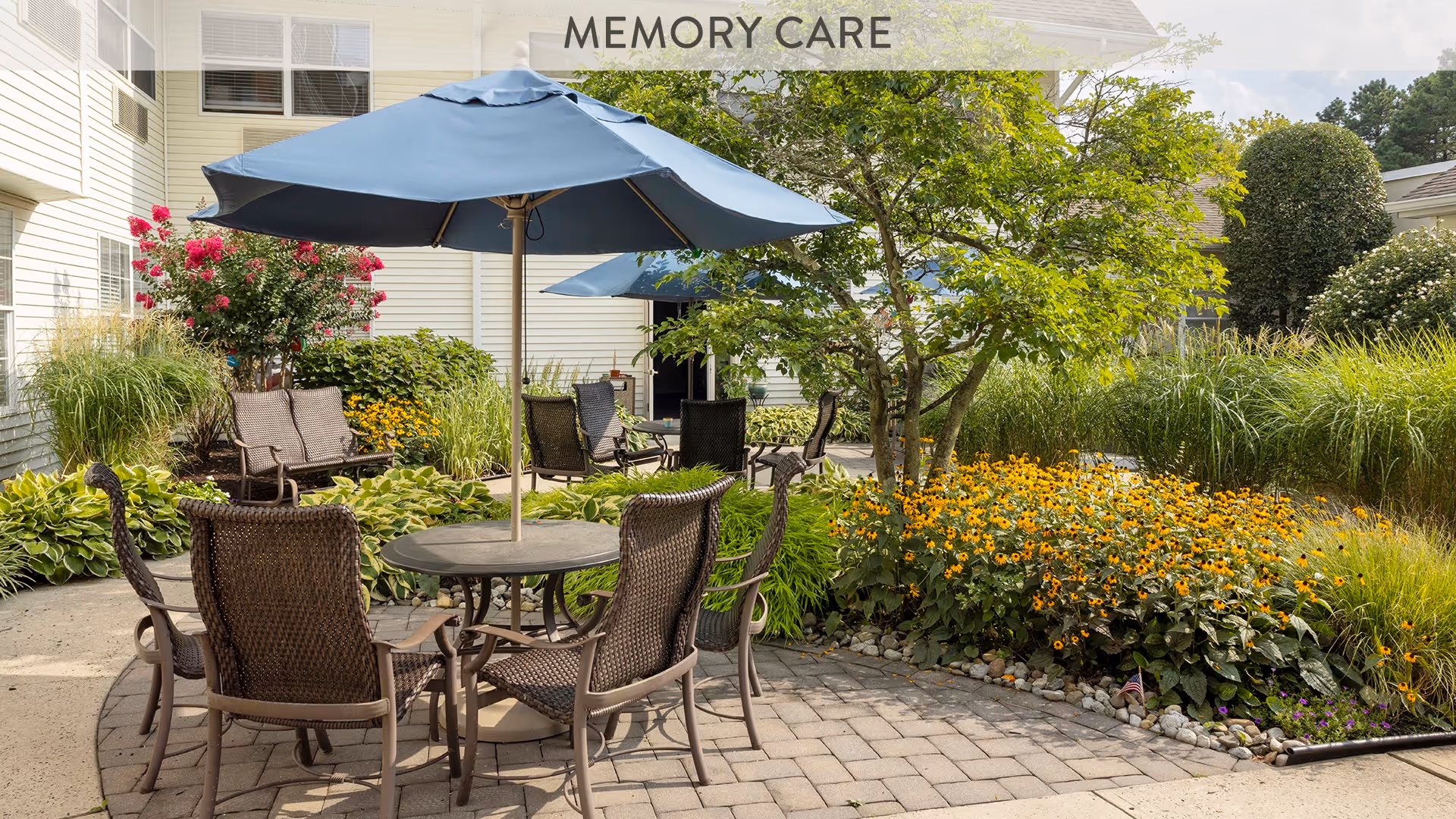 Outdoor patio area with round tables and wicker chairs under blue umbrellas, surrounded by lush greenery and colorful flowers in a garden setting at a memory care facility.