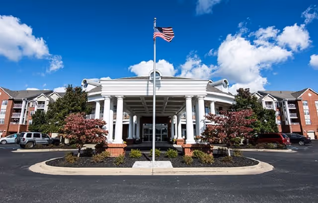 Front exterior view of Elison Independent & Assisted Living of Maplewood building with a covered entrance supported by white columns, an American flag on a flagpole in front, landscaped bushes and trees, and parked cars on either side under a blue sky with clouds.