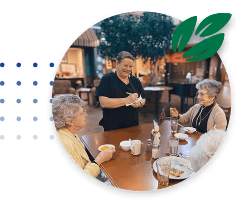 A smiling waitress takes an order from three elderly women seated around a dining table in a senior living facility. The table has cups, glasses, and plates with food. The background shows a warm, inviting dining area with tables and chairs.