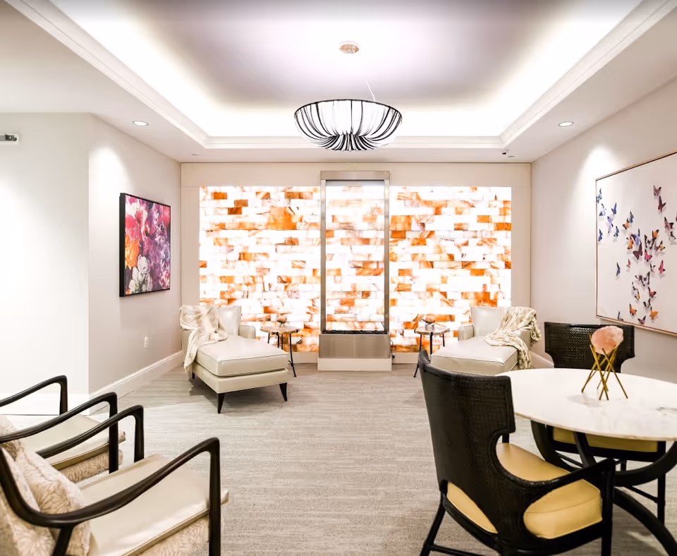 A bright and modern lounge area with two white chaise lounges draped with light blankets, two black armchairs with beige cushions, and a round marble table with a small floral centerpiece. The back wall features a decorative illuminated panel with a warm orange and white pattern, flanked by two small side tables. The ceiling has a recessed light fixture with a stylish black and white chandelier. Colorful artwork decorates the side walls.