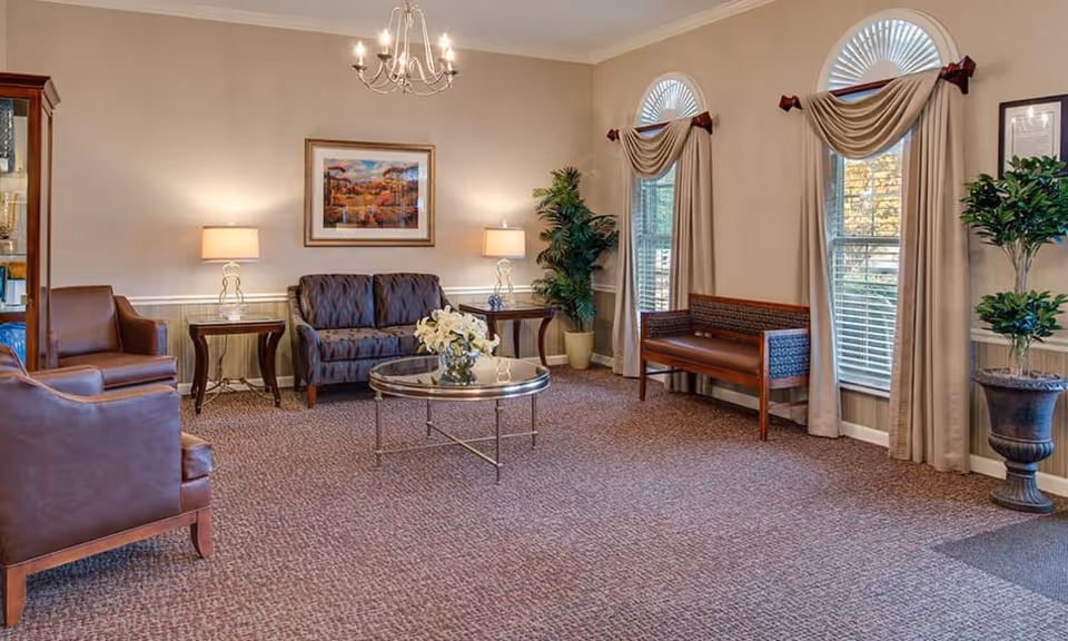 A cozy living room area with a patterned carpet, two leather armchairs, a patterned loveseat, a wooden bench, and a round glass coffee table with a flower arrangement. The room has beige walls, three tall windows with beige curtains and valances, two table lamps on side tables, a chandelier, framed artwork on the wall, and two potted plants.