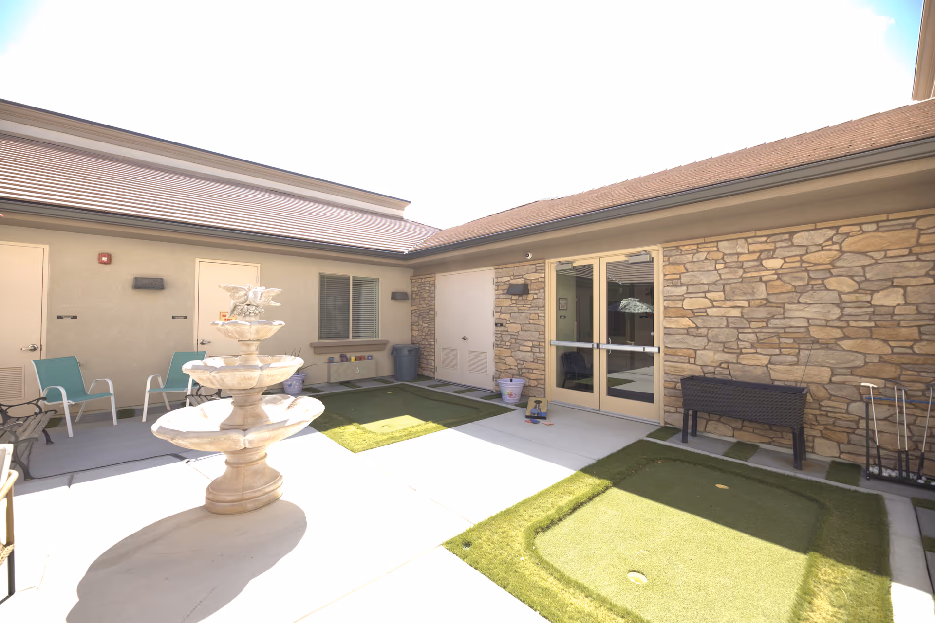 Outdoor courtyard area with a three-tier stone fountain in the center, surrounded by a concrete patio and artificial grass patches. There are two teal chairs against a beige wall with windows and doors, and a stone wall with double glass doors. Various small items and planters are placed around the area.