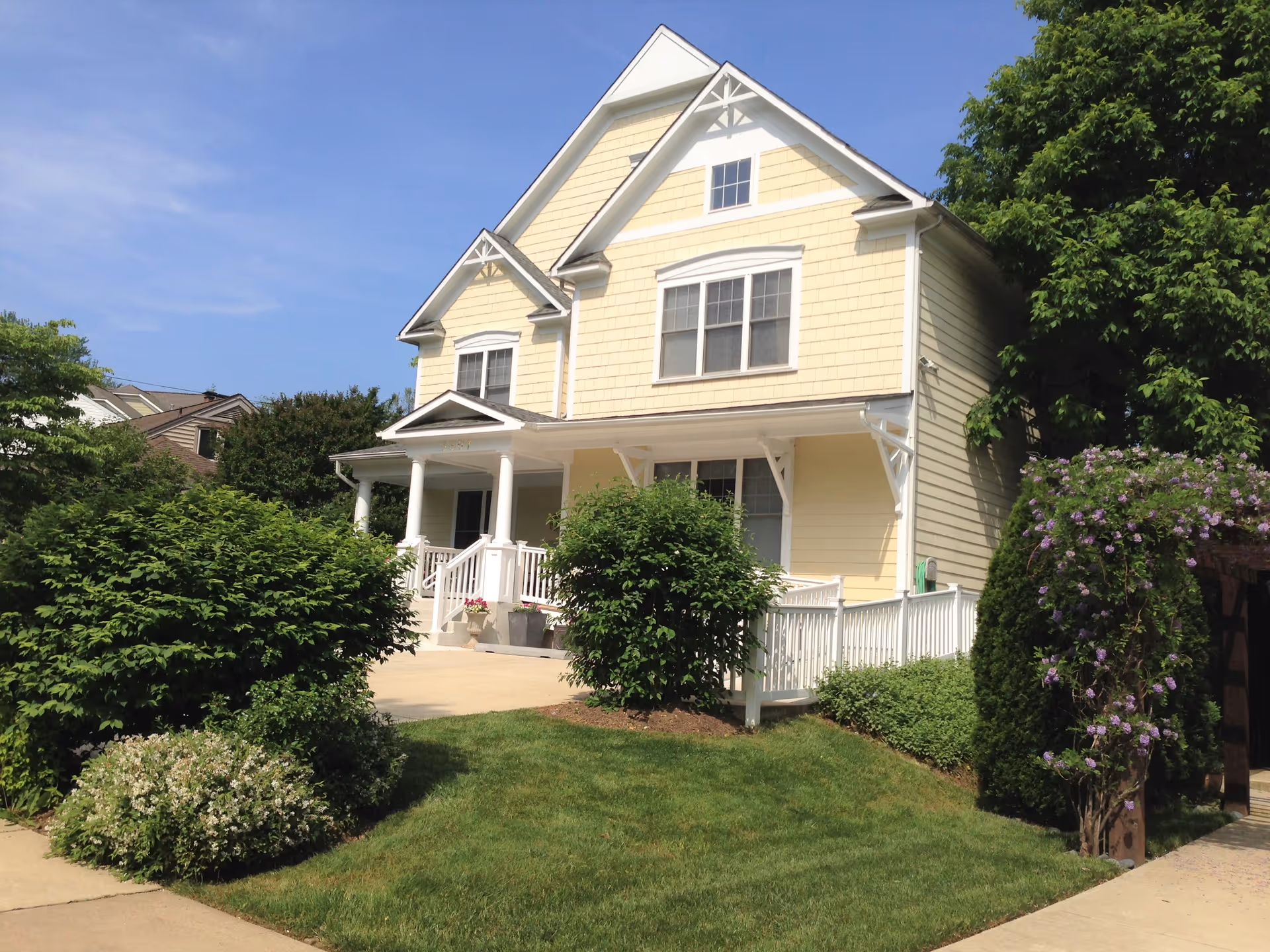 Two-story yellow house with white trim and a front porch, surrounded by shrubs and a lawn.