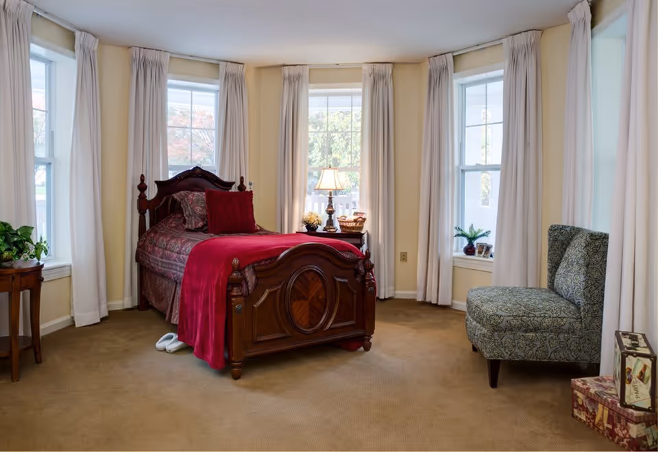A cozy bedroom with a wooden bed featuring a red blanket and pillows. The room has beige walls and carpet, three large windows with white curtains, a small wooden side table with a plant, a nightstand with a lamp and decorative items, a patterned upholstered chair, and two decorative suitcases stacked in the corner.
