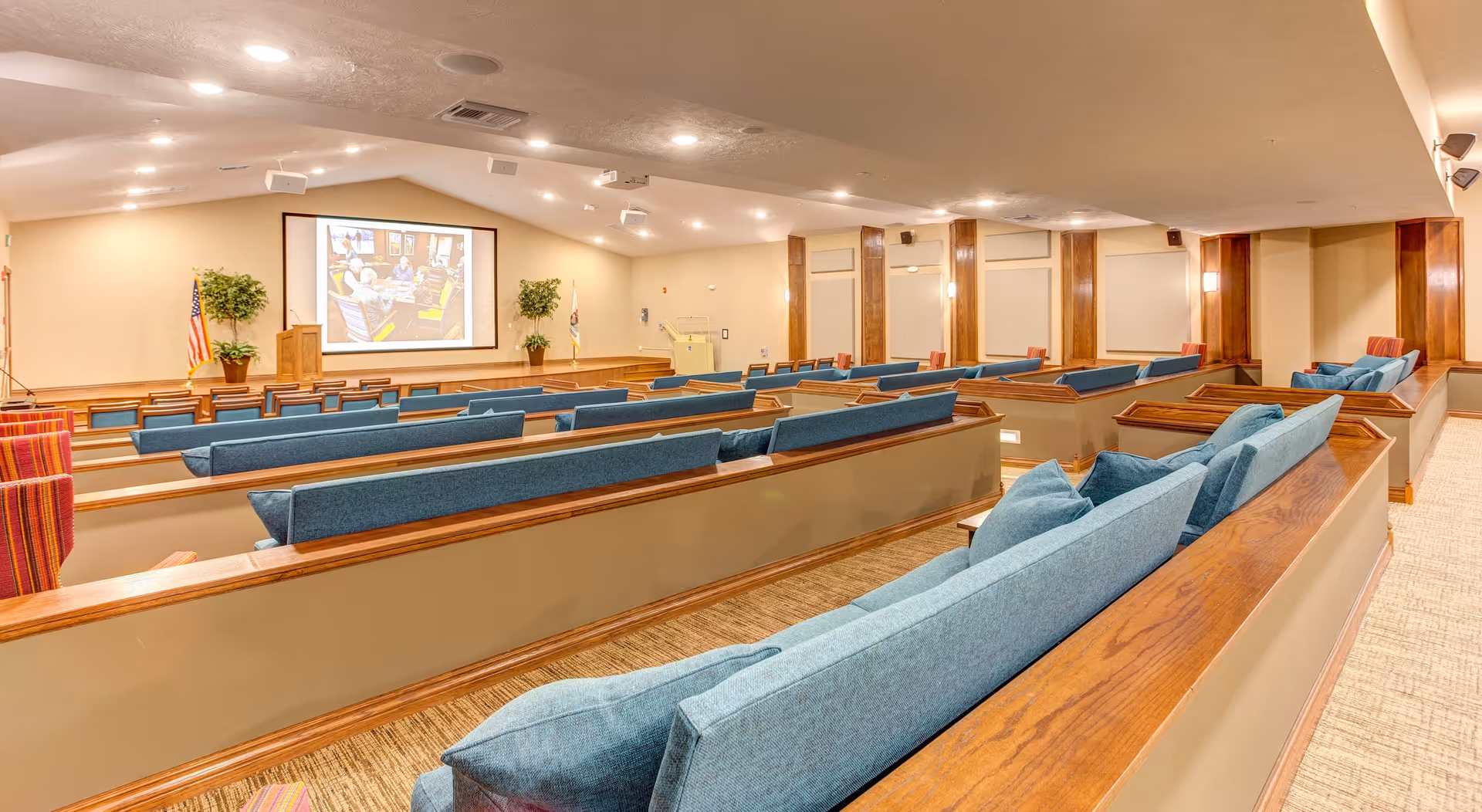 A spacious auditorium or presentation room with tiered seating featuring blue cushioned benches and wooden armrests. At the front, there is a large projection screen displaying an image, flanked by two potted plants and an American flag. The room has beige walls, recessed lighting in the ceiling, and carpeted floors.