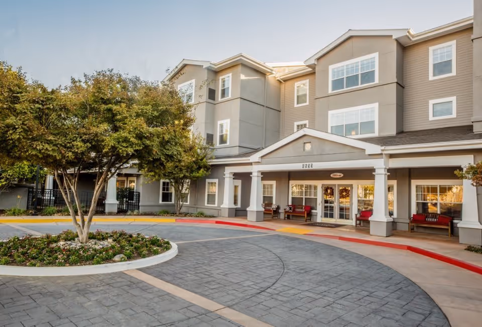 Exterior view of a three-story senior living facility with beige and gray walls, white trim, and multiple windows. The entrance features a covered porch with white columns and benches with red cushions. A circular driveway with a landscaped island containing a tree and flowers is in front of the building.