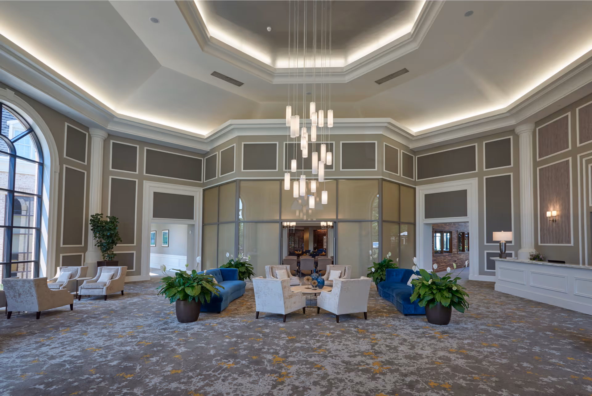 Spacious and elegant senior living community lounge with high ceilings, large windows, modern hanging light fixtures, blue sofas, beige armchairs arranged around coffee tables, potted plants, and a reception desk on the right side.