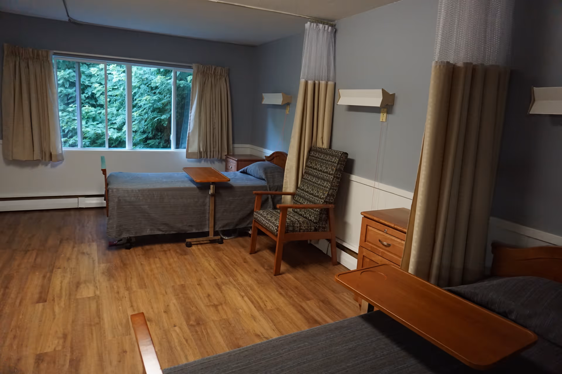 A nursing home bedroom with two beds, each equipped with a wooden overbed table. The room has wood flooring, light blue walls with white trim, beige curtains on a large window showing green foliage outside, and a patterned armchair between the beds. Privacy curtains are partially drawn around each bed.
