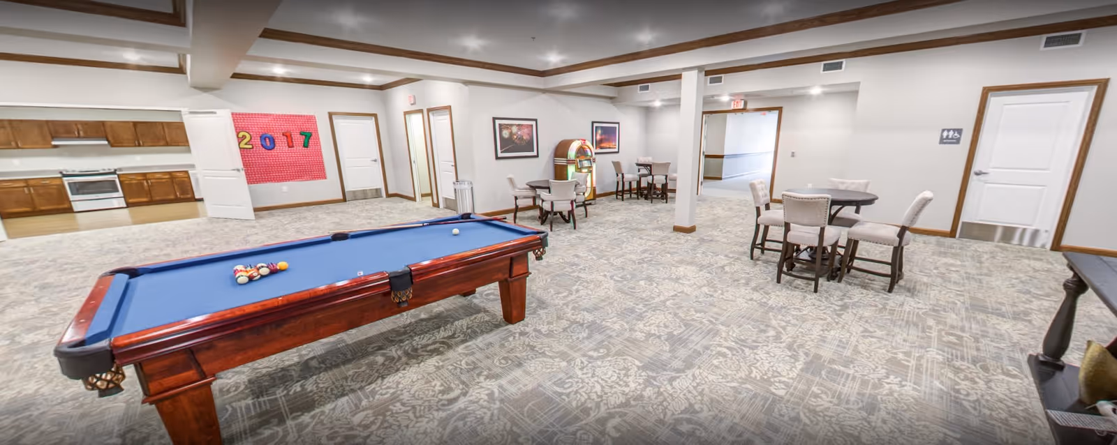 A spacious senior living common area featuring a pool table with balls arranged for a game, several small round tables with upholstered chairs, a jukebox, and a kitchenette with wooden cabinets and a stove. The room has patterned carpet flooring, white walls with wooden trim, and multiple doors including one marked with a restroom sign.