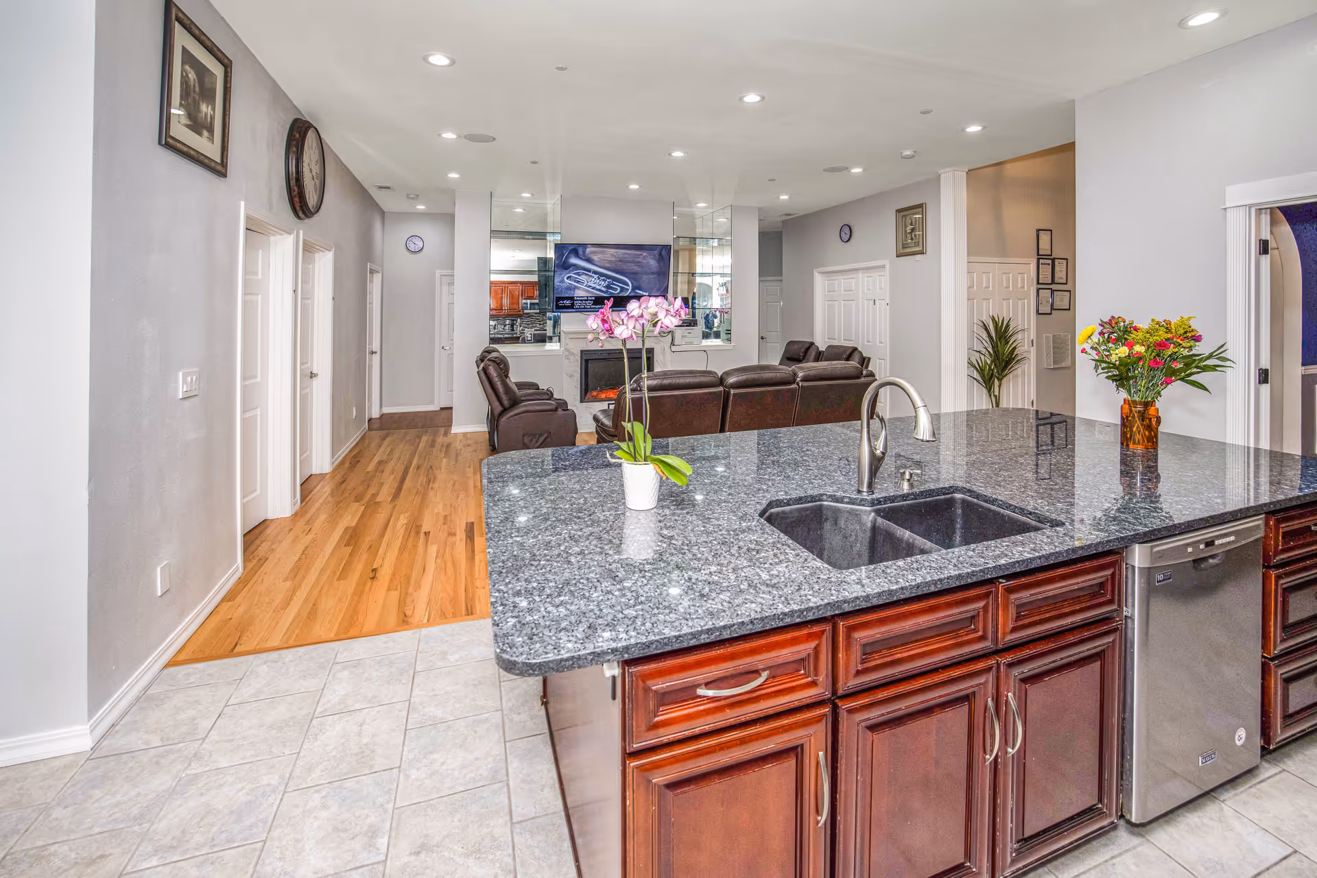 Interior view of a modern living space featuring a kitchen island with a granite countertop, a double sink, and a dishwasher. The island has dark wooden cabinets and is decorated with a small potted orchid and a vase of colorful flowers. Beyond the kitchen is a living room area with brown leather recliners facing a wall-mounted TV above a fireplace. The floor transitions from tiled in the kitchen to hardwood in the living area. The walls are light gray with framed pictures and clocks, and there are several white doors along the hallway.
