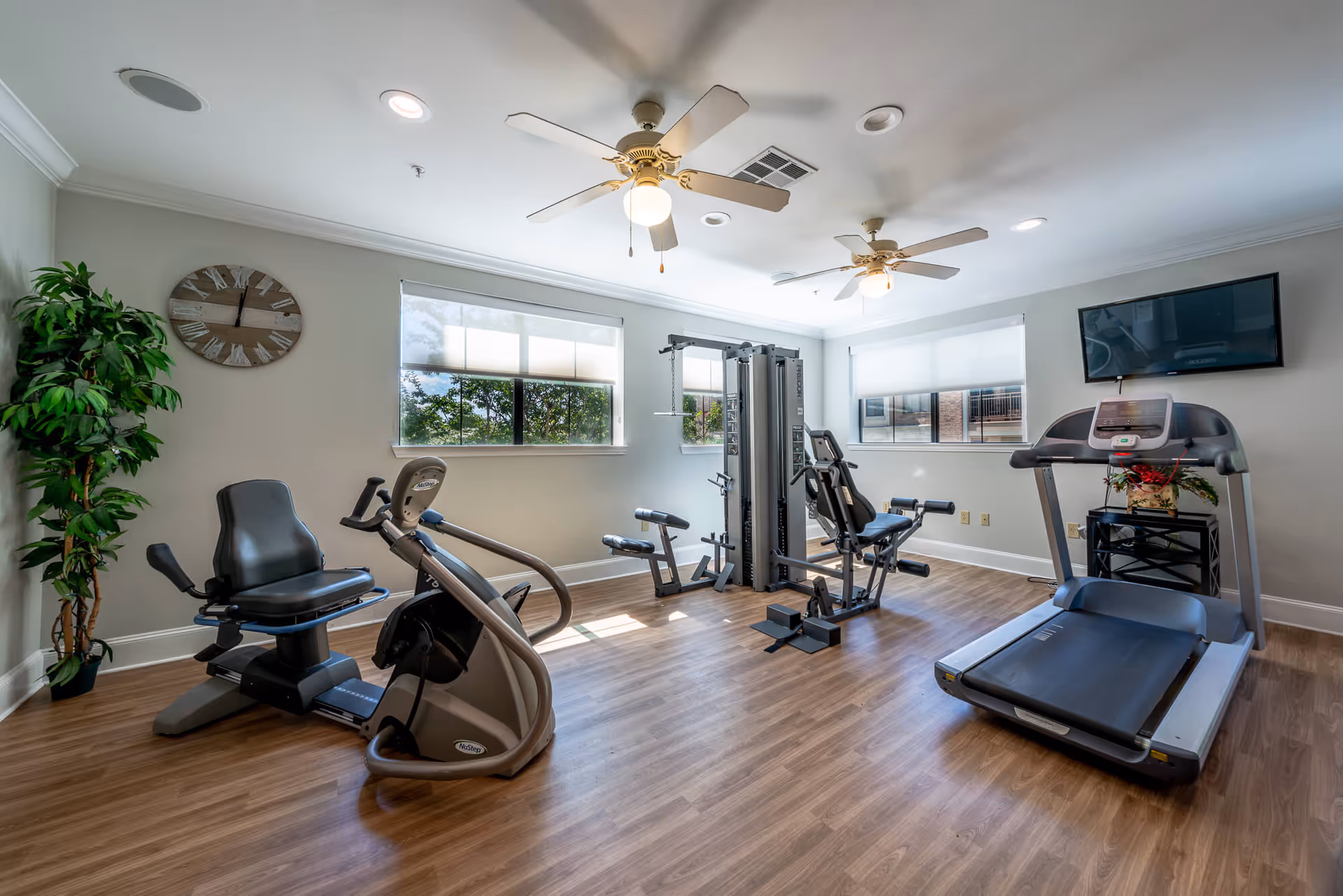 A bright fitness room with wooden flooring featuring exercise equipment including a recumbent bike, a multi-station gym machine, and a treadmill. The room has two ceiling fans with lights, a wall-mounted TV, two windows with white blinds, a large wall clock, and a potted plant in the corner.
