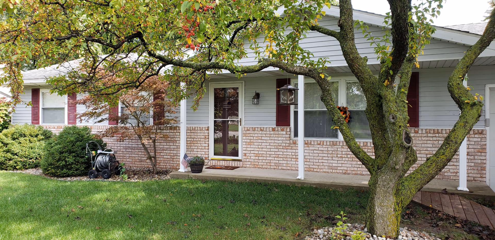 Front exterior view of a single-story house with light gray siding and brick accents. There is a covered porch with a glass storm door, a small American flag in a planter, and a tree with green leaves in the foreground. Bushes and a garden hose reel are visible near the house.