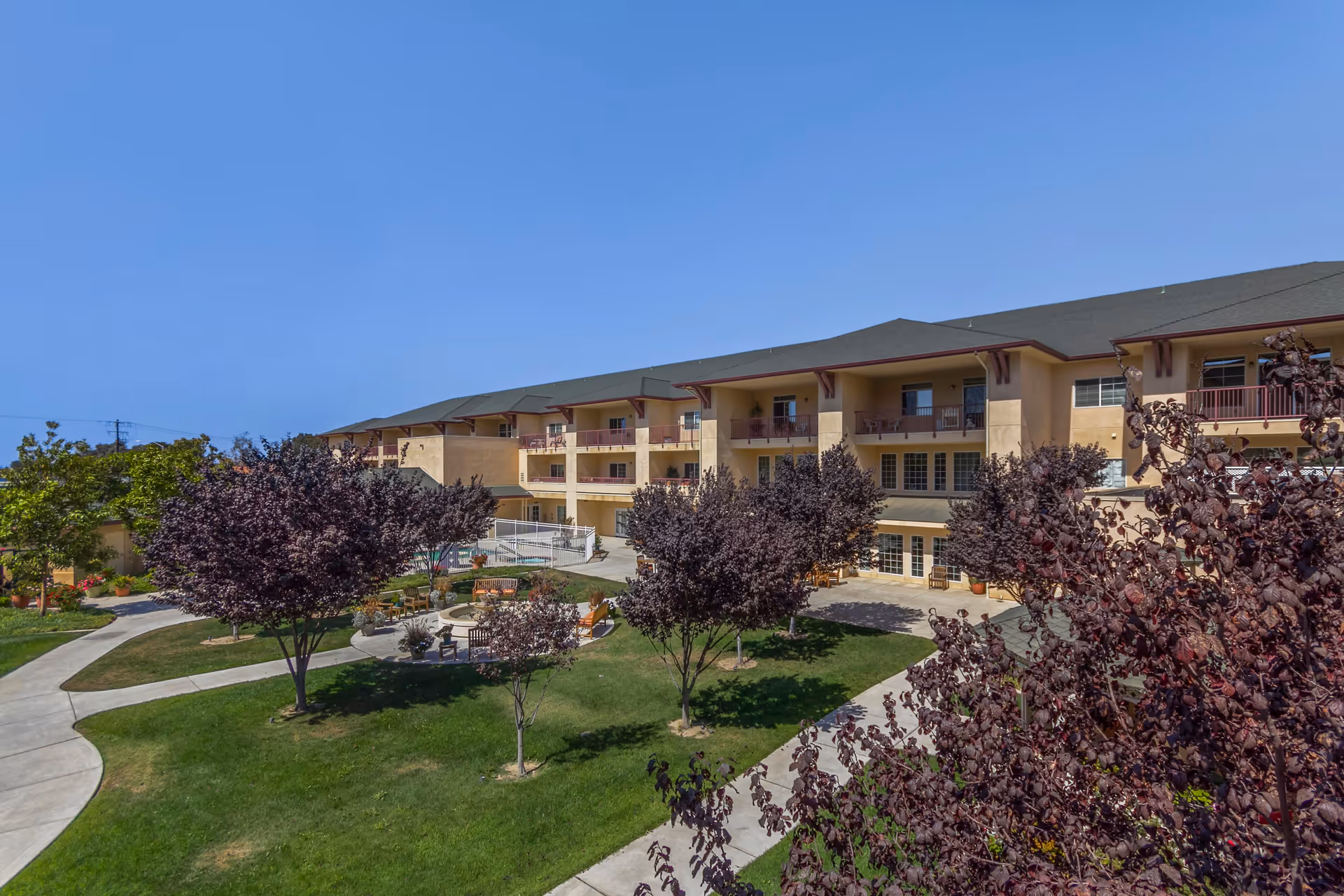 Outdoor view of a senior living facility with a three-story beige building in the background, surrounded by a well-maintained garden area with green grass, several trees with dark purple leaves, paved walkways, and outdoor seating areas under a clear blue sky.