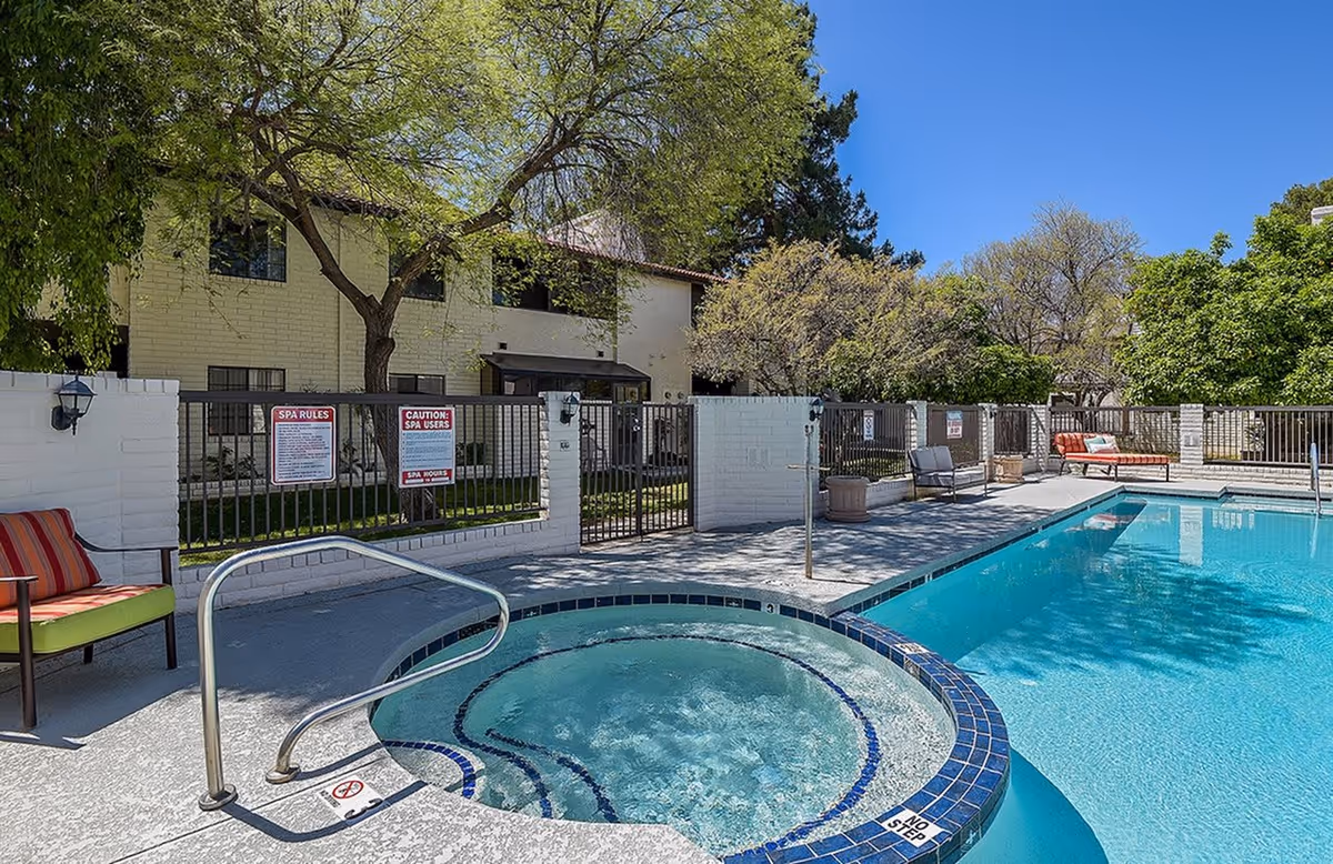Outdoor pool area at Zen Senior Living featuring a round hot tub with bubbling water in the foreground and a rectangular swimming pool beside it. There are several chairs with cushions around the pool area, a white brick fence with metal gates, and trees providing shade. The sky is clear and blue.