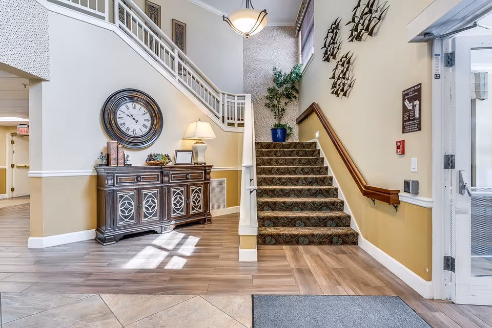 Interior view of a senior living facility entrance area featuring a staircase with patterned carpet, a wooden sideboard with decorative items and a large round wall clock above it, a potted plant at the top of the stairs, and a hanging light fixture. The walls are painted beige and white, and there is a glass door on the right side.