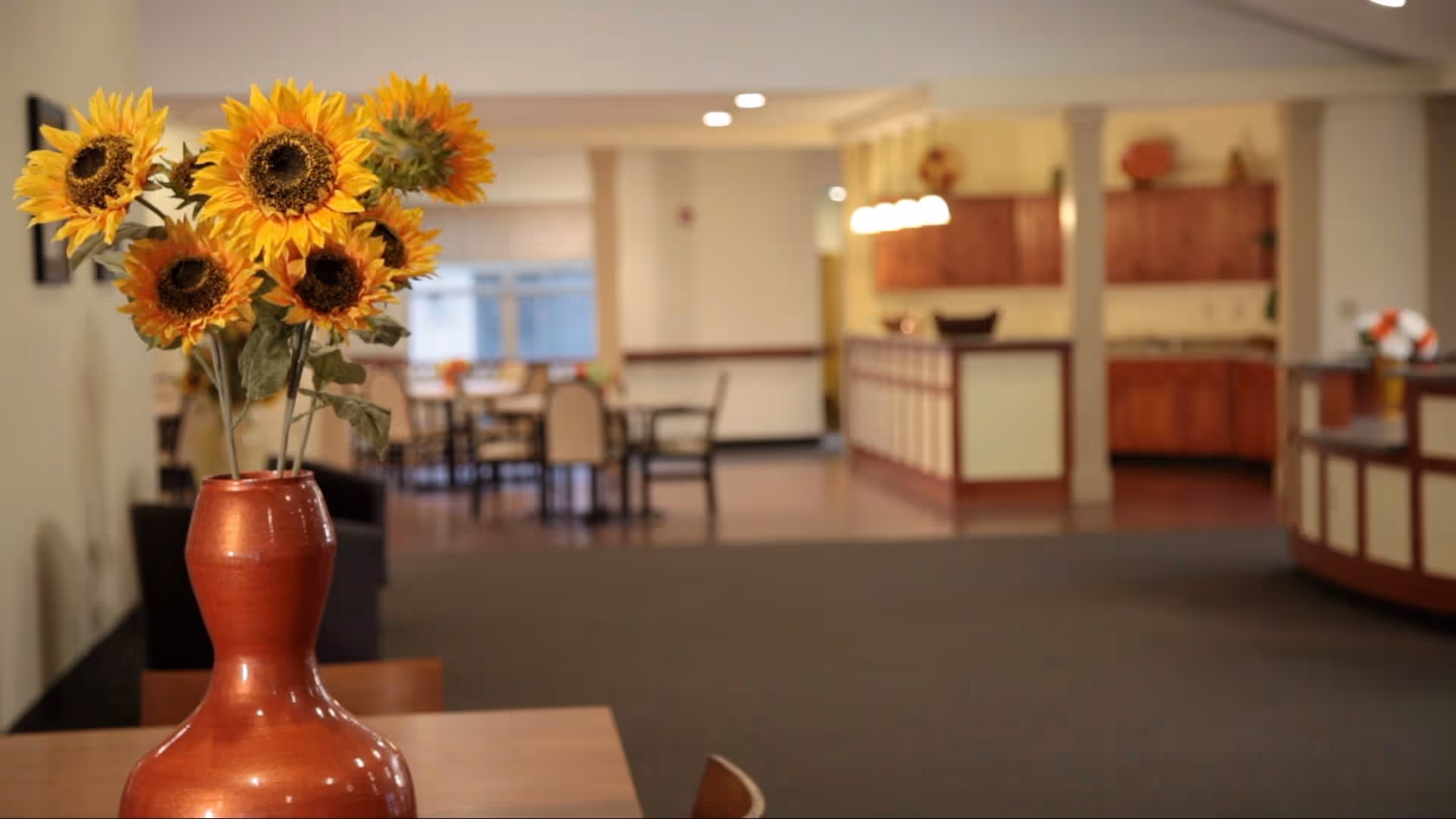 A close-up of a red vase with bright yellow sunflowers on a wooden table in a well-lit room. In the background, there are dining tables and chairs, and a kitchen area with wooden cabinets and hanging lights.