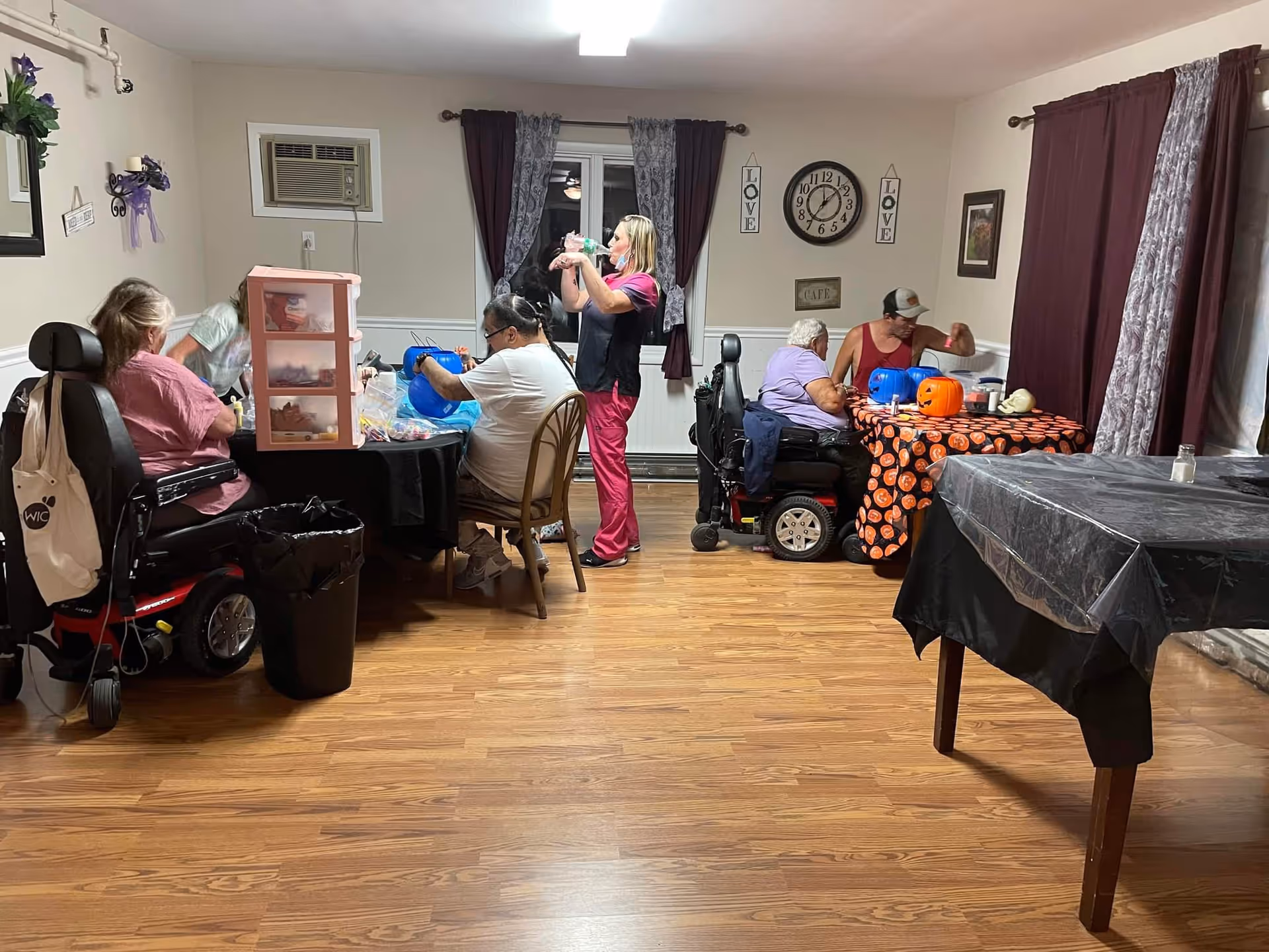 A group of elderly individuals and a caregiver are gathered in a room with wooden flooring and beige walls. Some of the elderly are seated in wheelchairs, while others sit on chairs around tables covered with Halloween-themed tablecloths and decorations. The caregiver is standing and interacting with the group. The room has dark curtains, a wall clock, and decorative signs that say 'LOVE'.