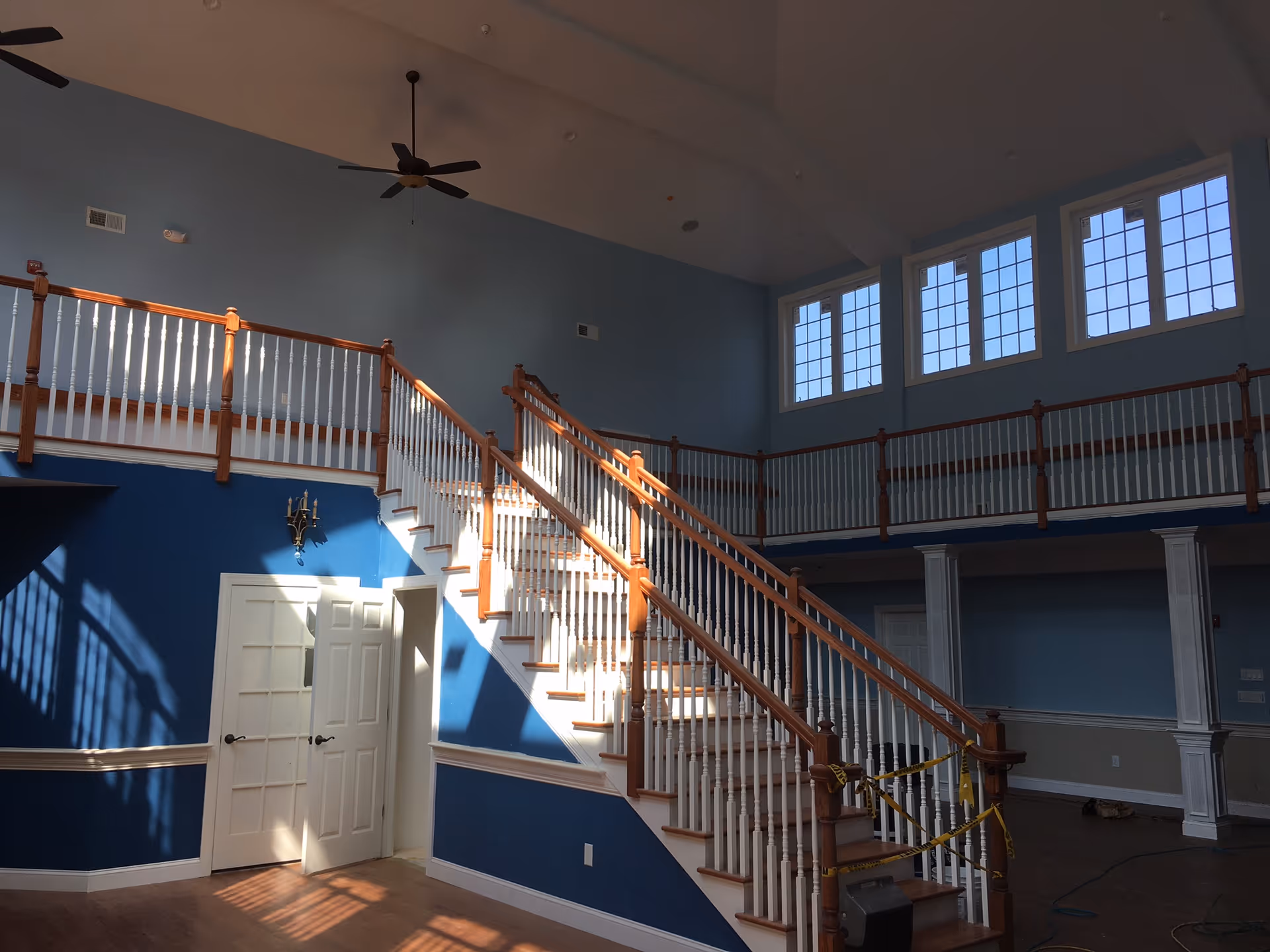 Interior view of a spacious area with a wooden staircase featuring white balusters and wooden handrails. The walls are painted blue with white trim, and there are large windows near the ceiling allowing natural light to illuminate the space. The upper level has a balcony with matching railing overlooking the lower floor. Ceiling fans are mounted on the high ceiling.