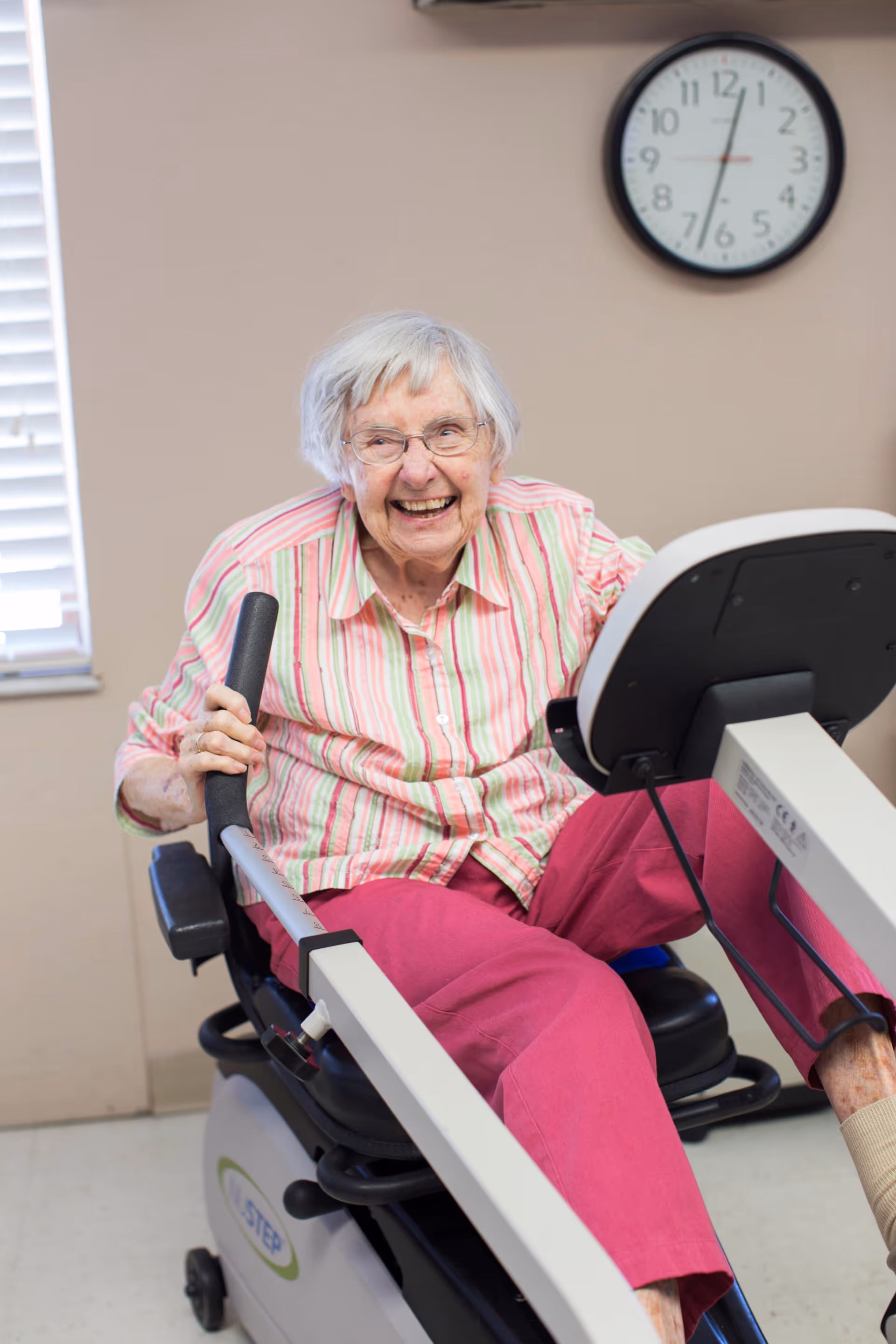 An elderly woman with white hair and glasses, wearing a striped shirt and pink pants, smiling while using a seated exercise machine in a room with a clock on the wall and a window with blinds.
